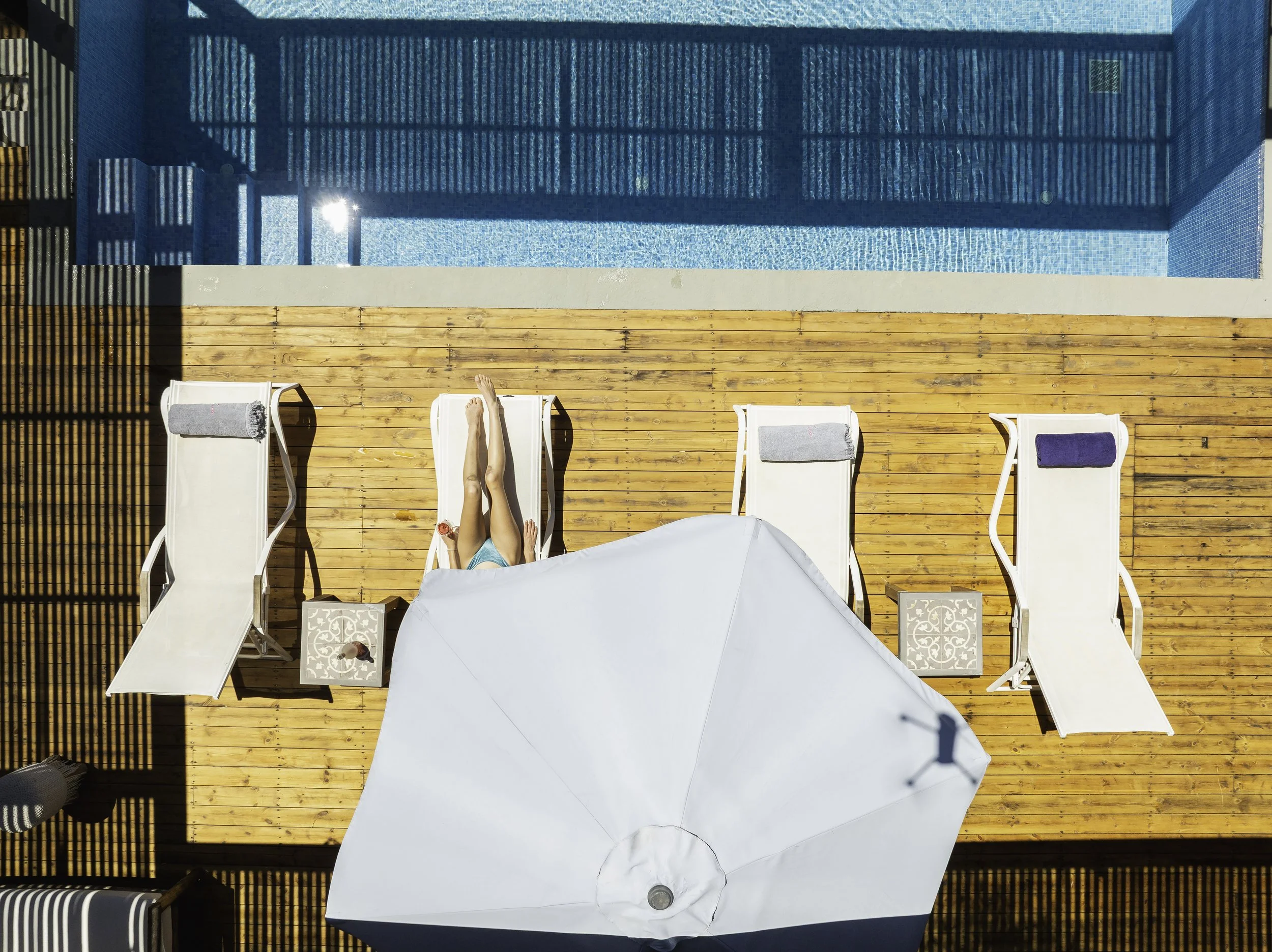 Top-down view of a swimming pool area with four lounge chairs on a wooden deck, an umbrella, and a person sunbathing.