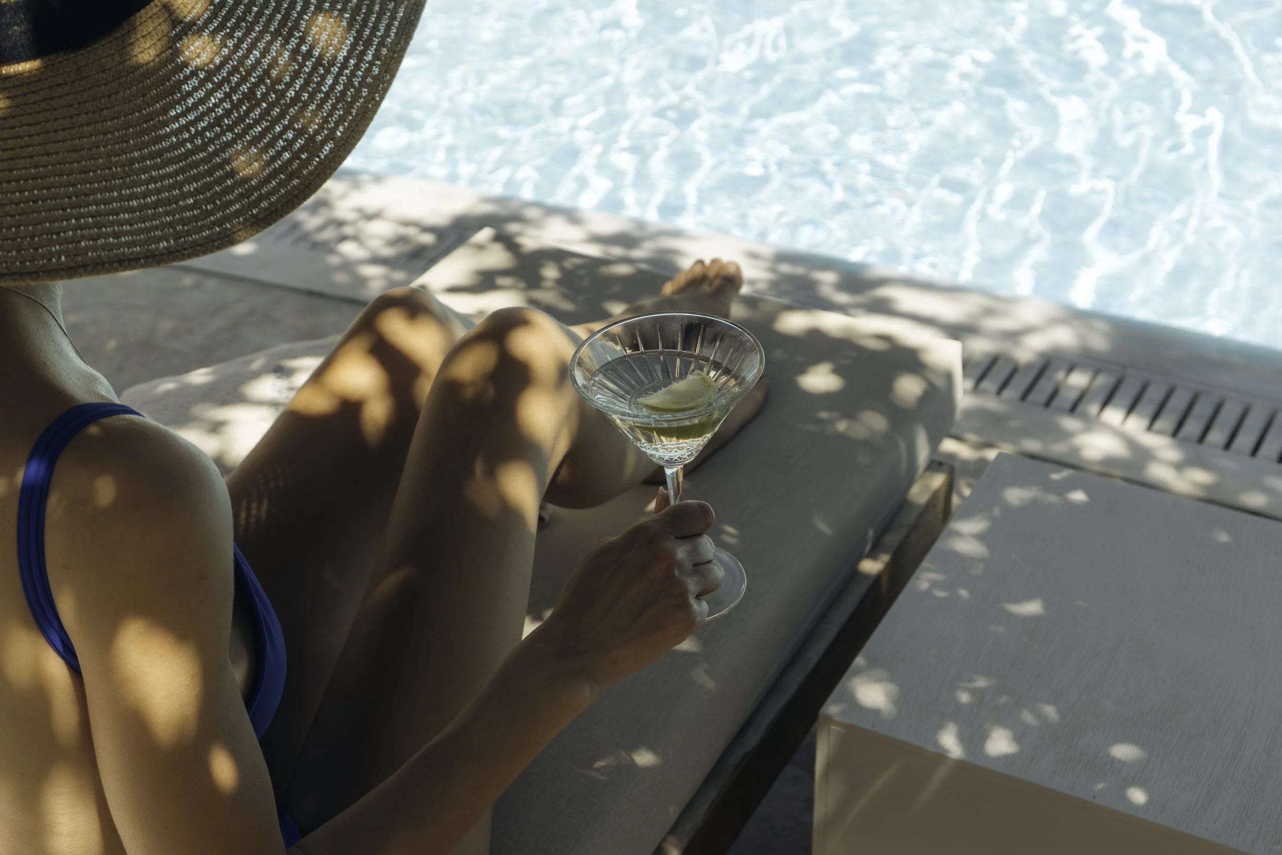 A woman relaxing on a lounge chair by the pool, wearing a large sunhat and holding a cocktail with lemon.
