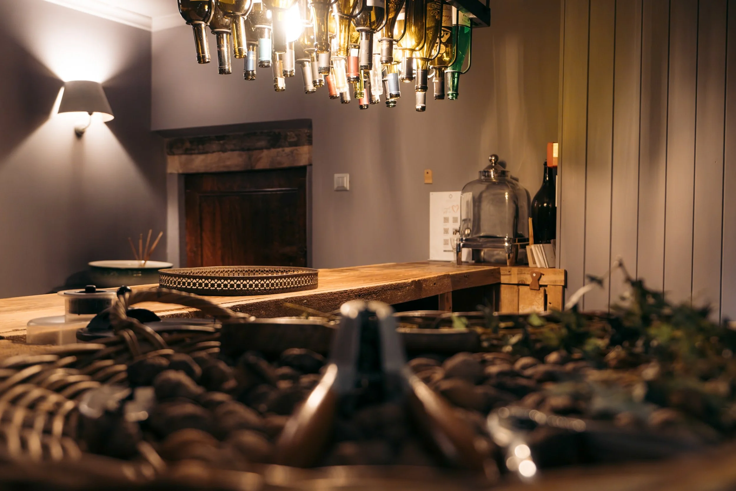 Interior of a cozy bar with a wooden countertop, hanging bottles of alcohol, and a modern light fixture. The scene shows a blurred basket with stones or decor in the foreground and warm lighting.