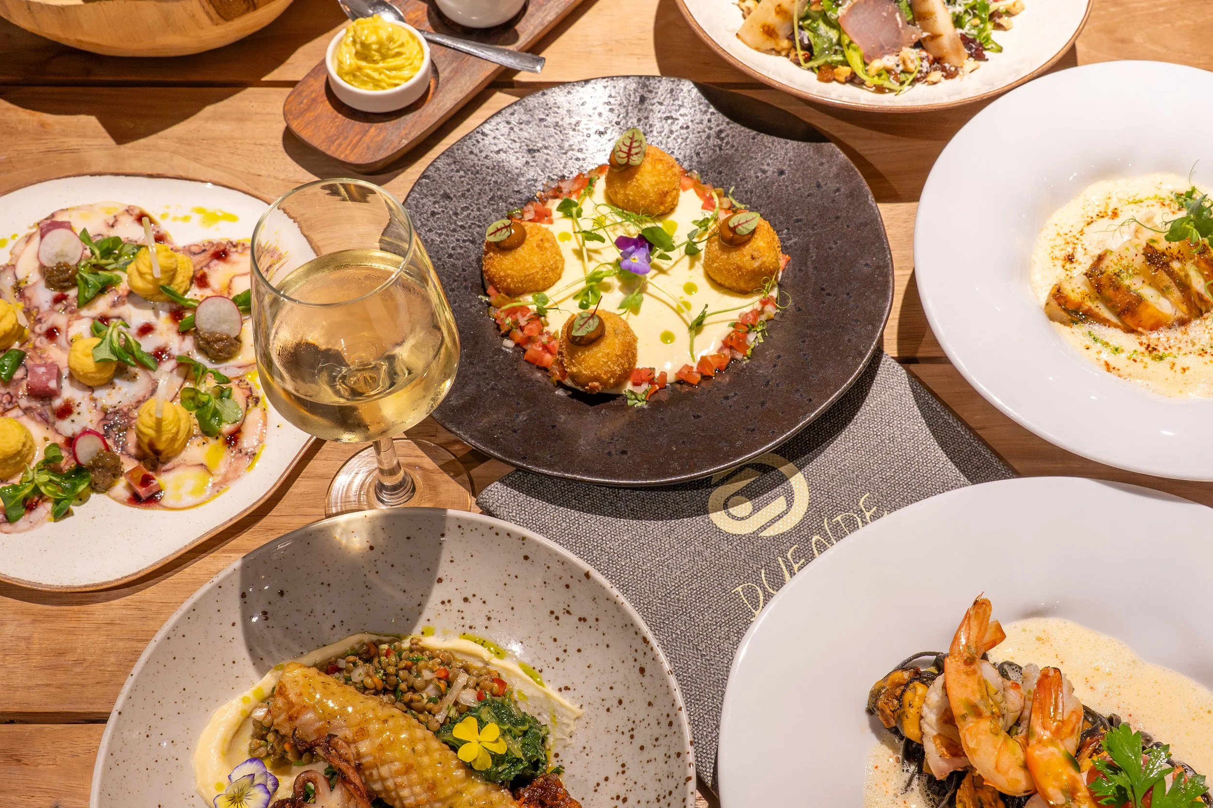 A table with assorted dishes including a white wine, seafood dishes, a salad, and appetizers, in a setting that suggests a restaurant meal.