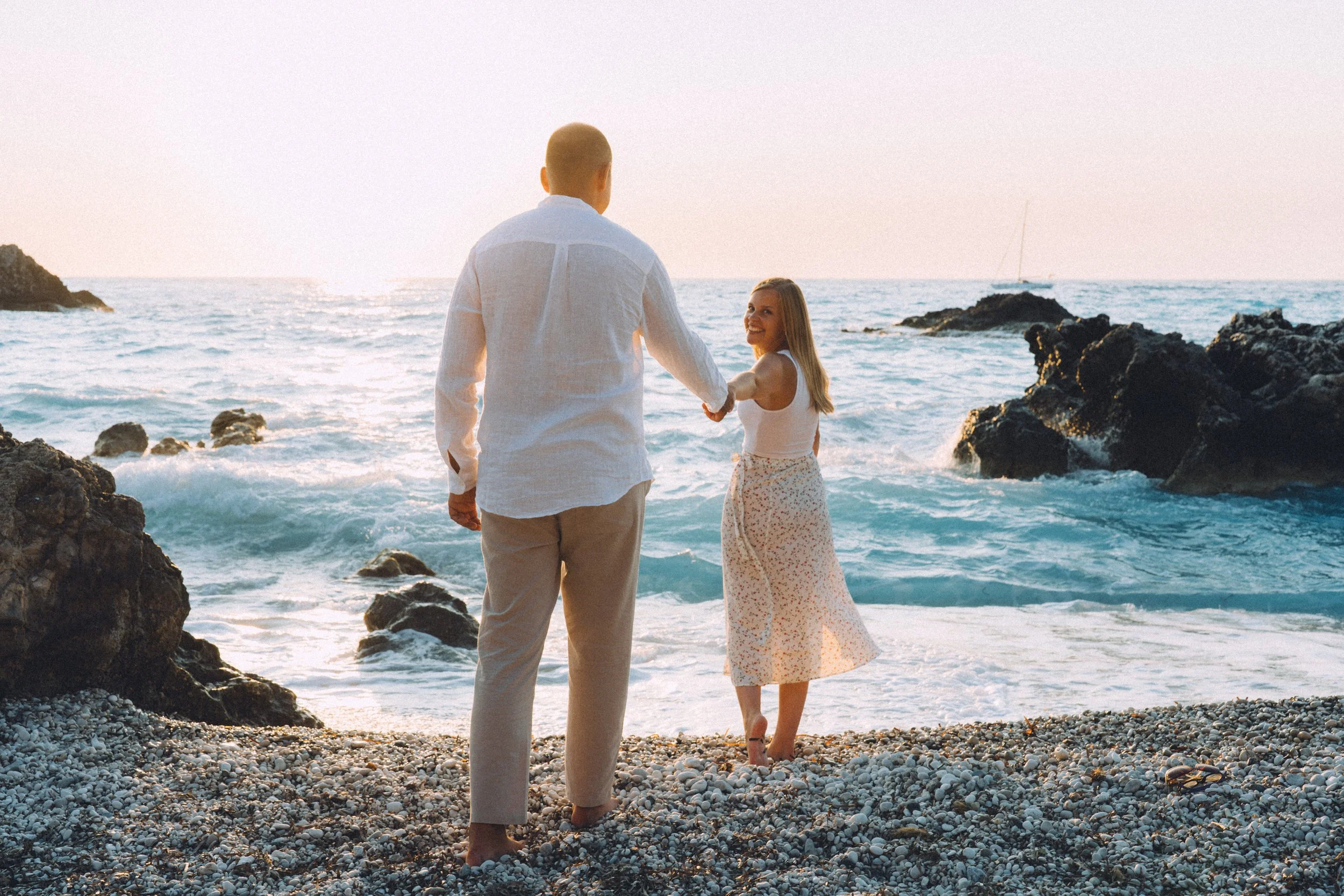 A man and woman holding hands on a rocky beach at sunset, with the ocean and sailboat in the background.