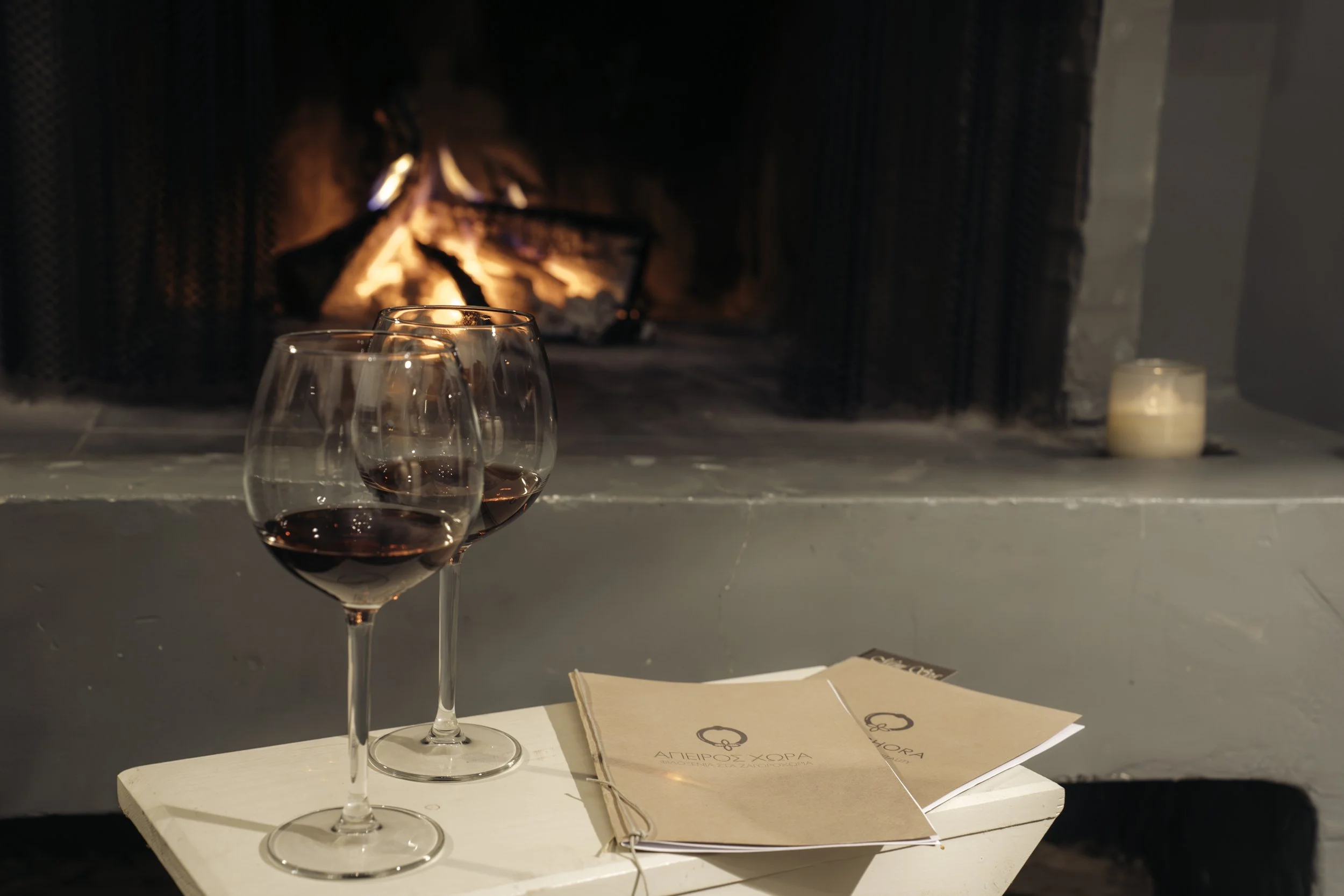 Two glasses of red wine on a small table in front of a fireplace, with a candle to the right and a menu on the table.