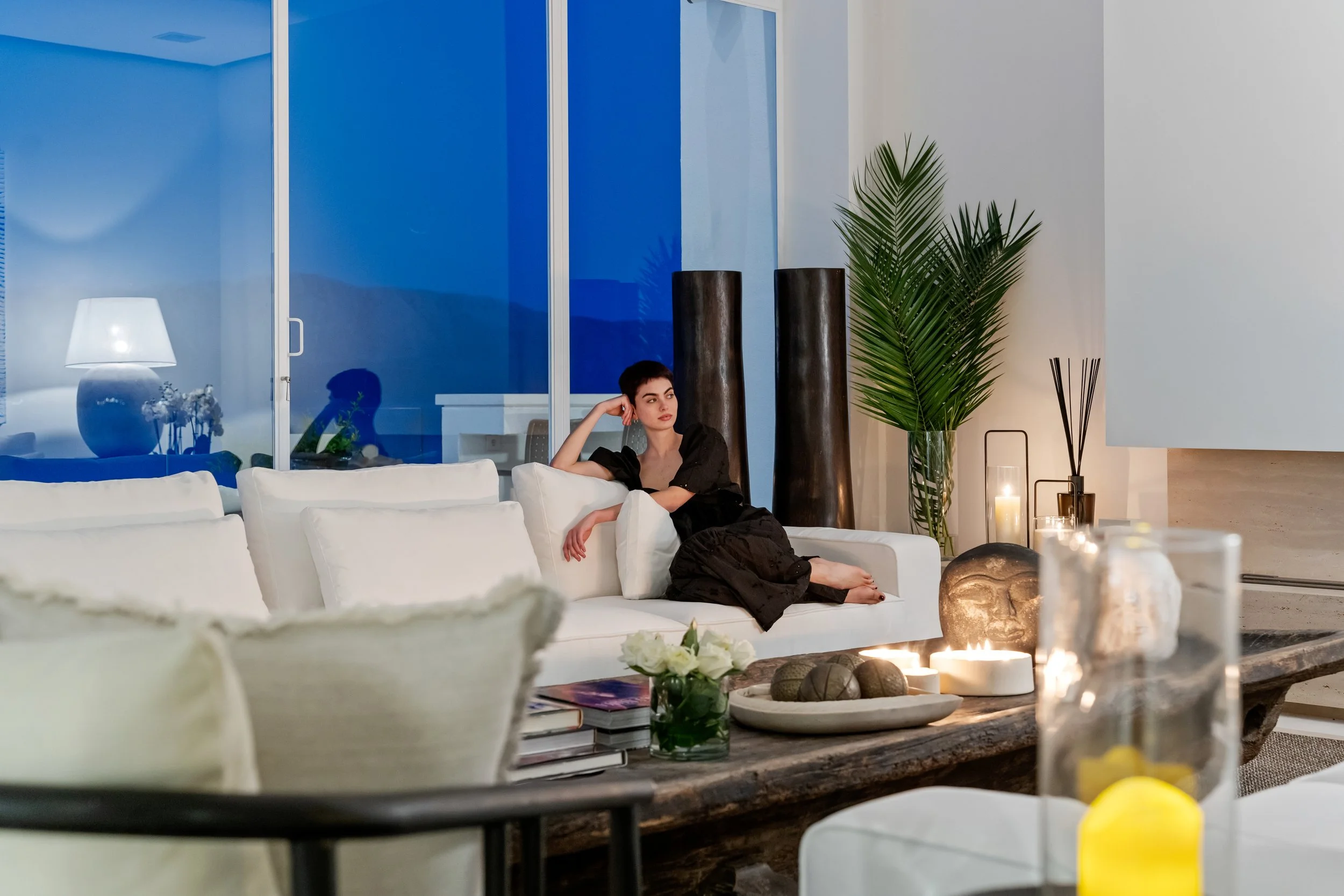 A woman in a black dress sitting on a white sofa in a modern living room with candles, plants, and decorative objects, during evening time.