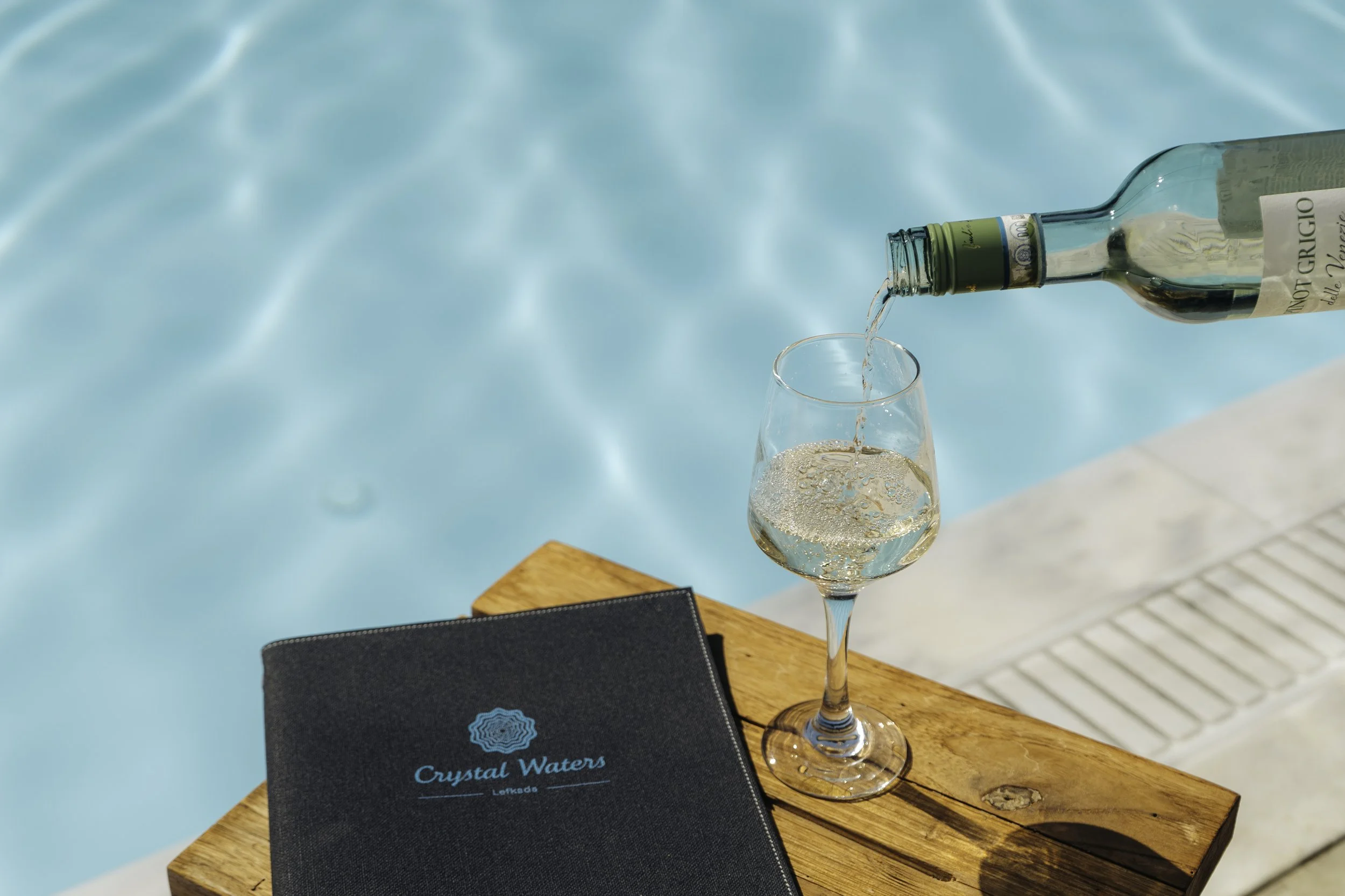 A glass of white wine on a wooden table near a swimming pool, with a bottle of wine pouring into the glass and a black booklet with the words "Crystal Waters" on it.