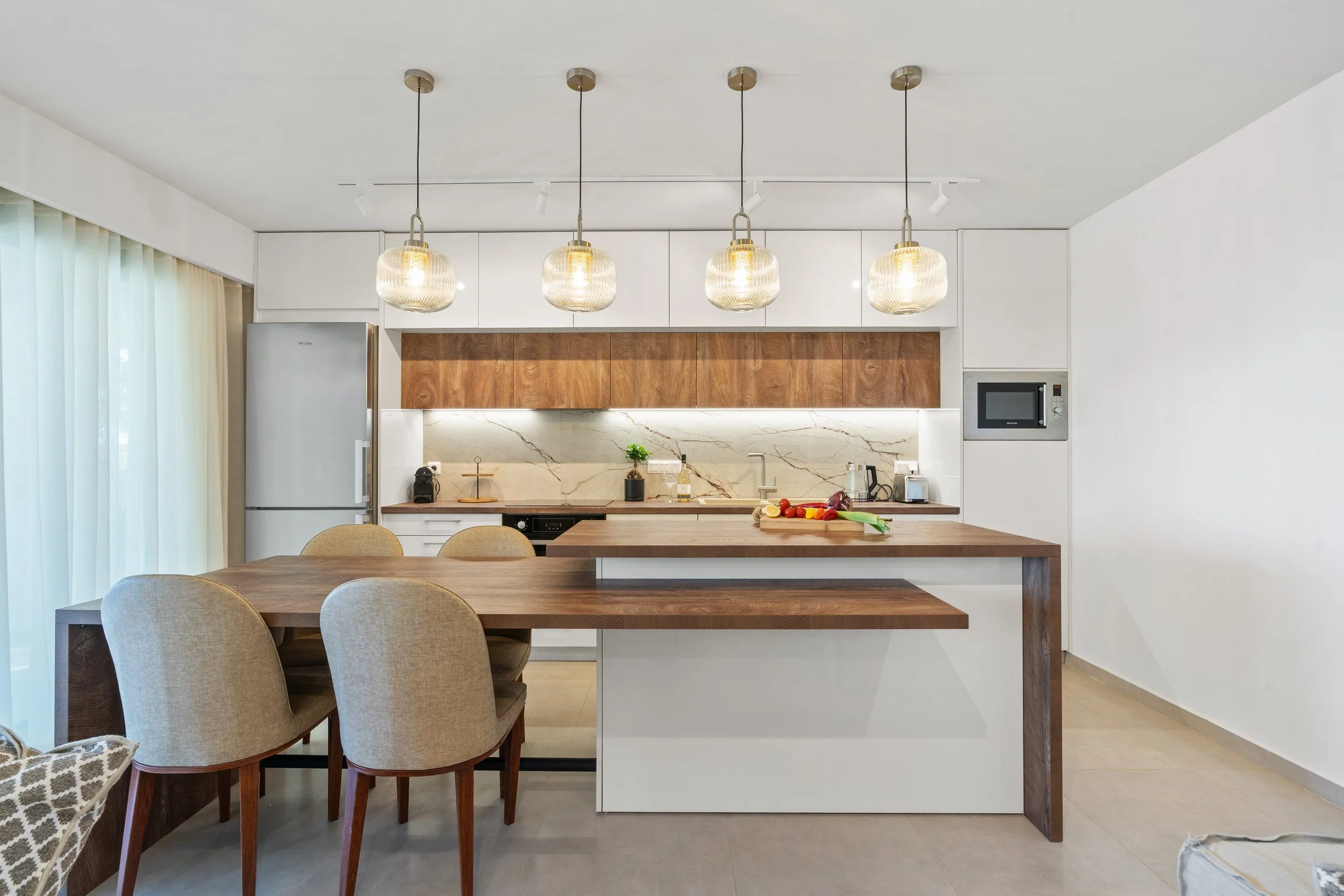 Modern kitchen with white cabinets, wooden accents, marble backsplash, and pendant lighting. Includes a dining table with chairs, a refrigerator, microwave, and fresh vegetables on the counter.