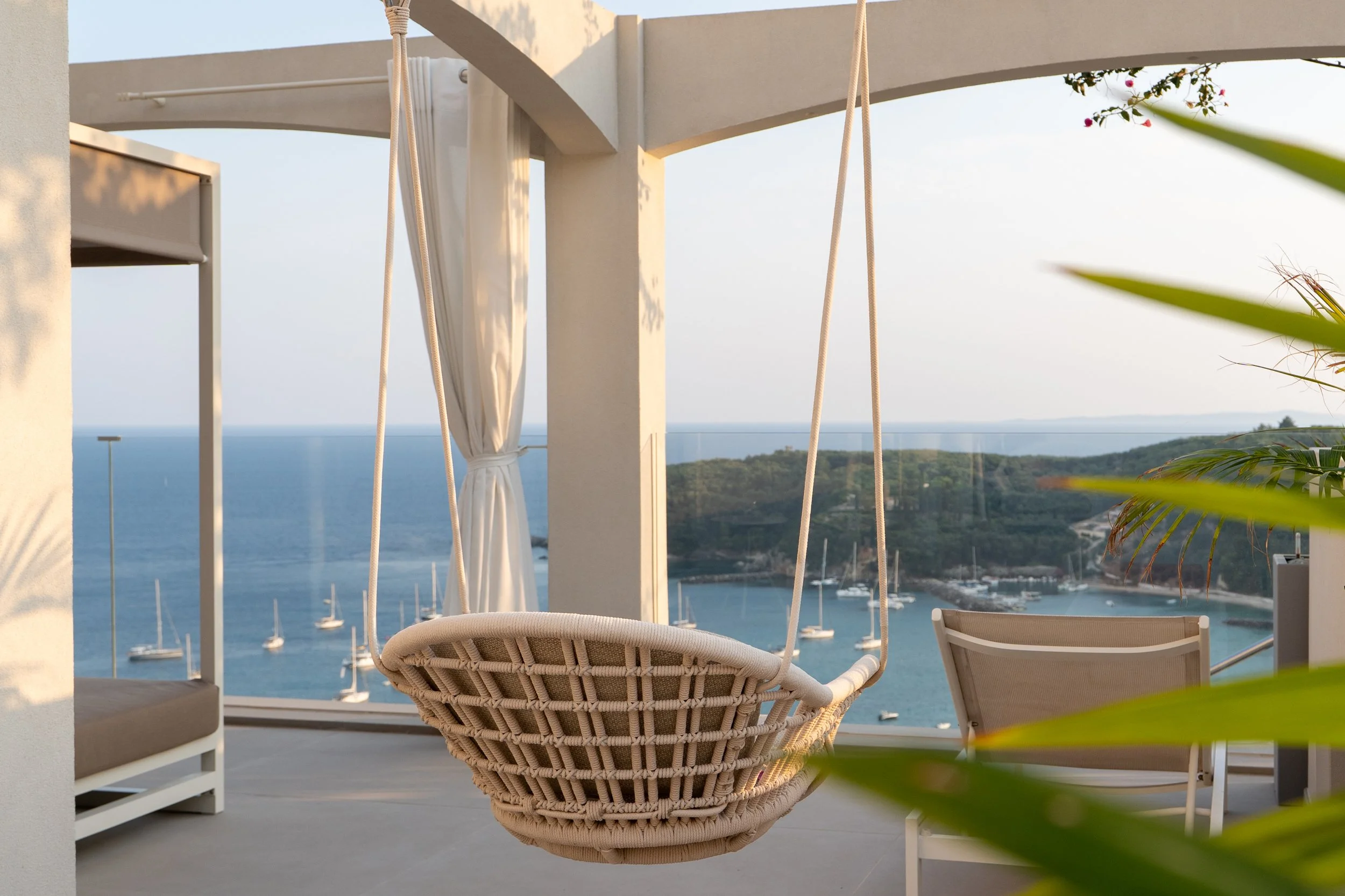 A balcony with a hanging chair overlooking a marina with boats, on a sunny day.