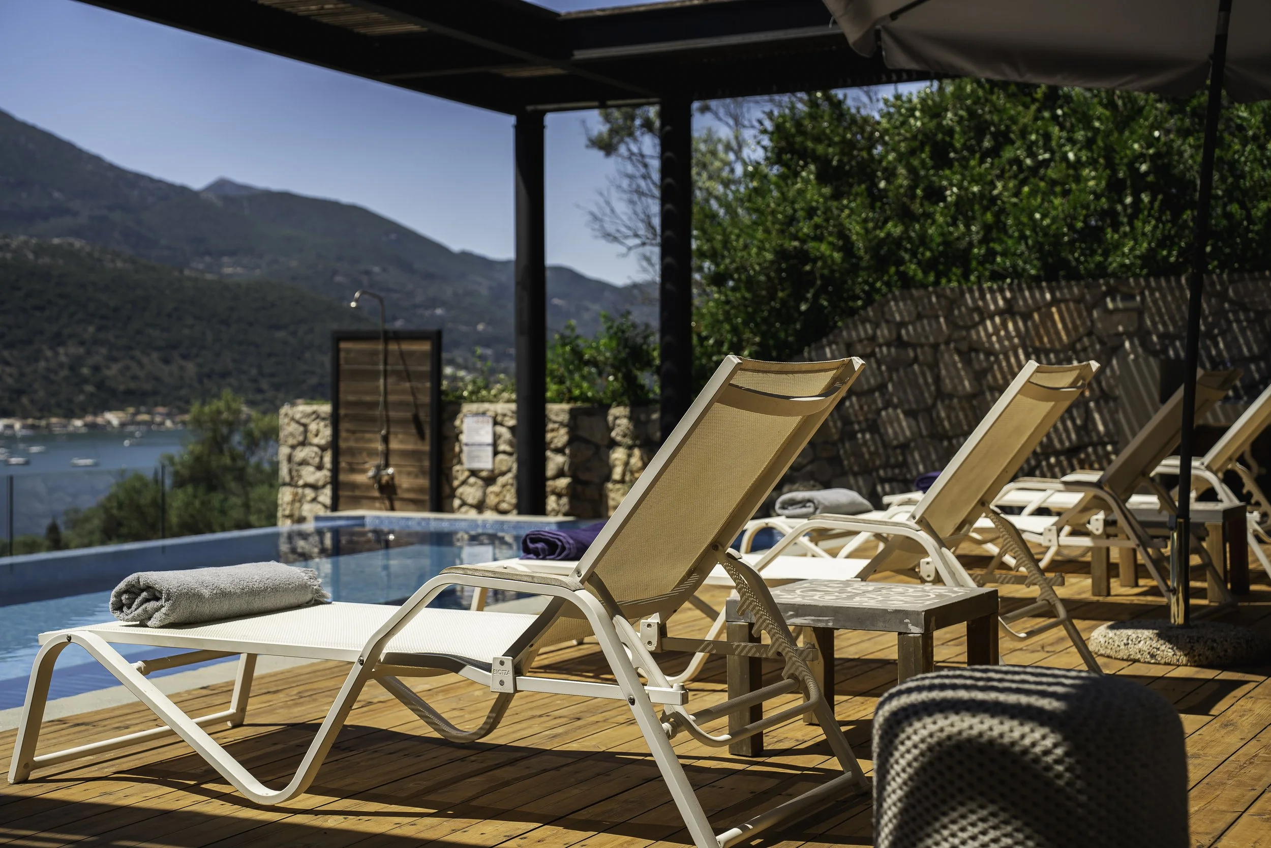 Sun loungers with towels near a swimming pool overlooking a mountain landscape and water, with a stone wall and outdoor shower area in the background.