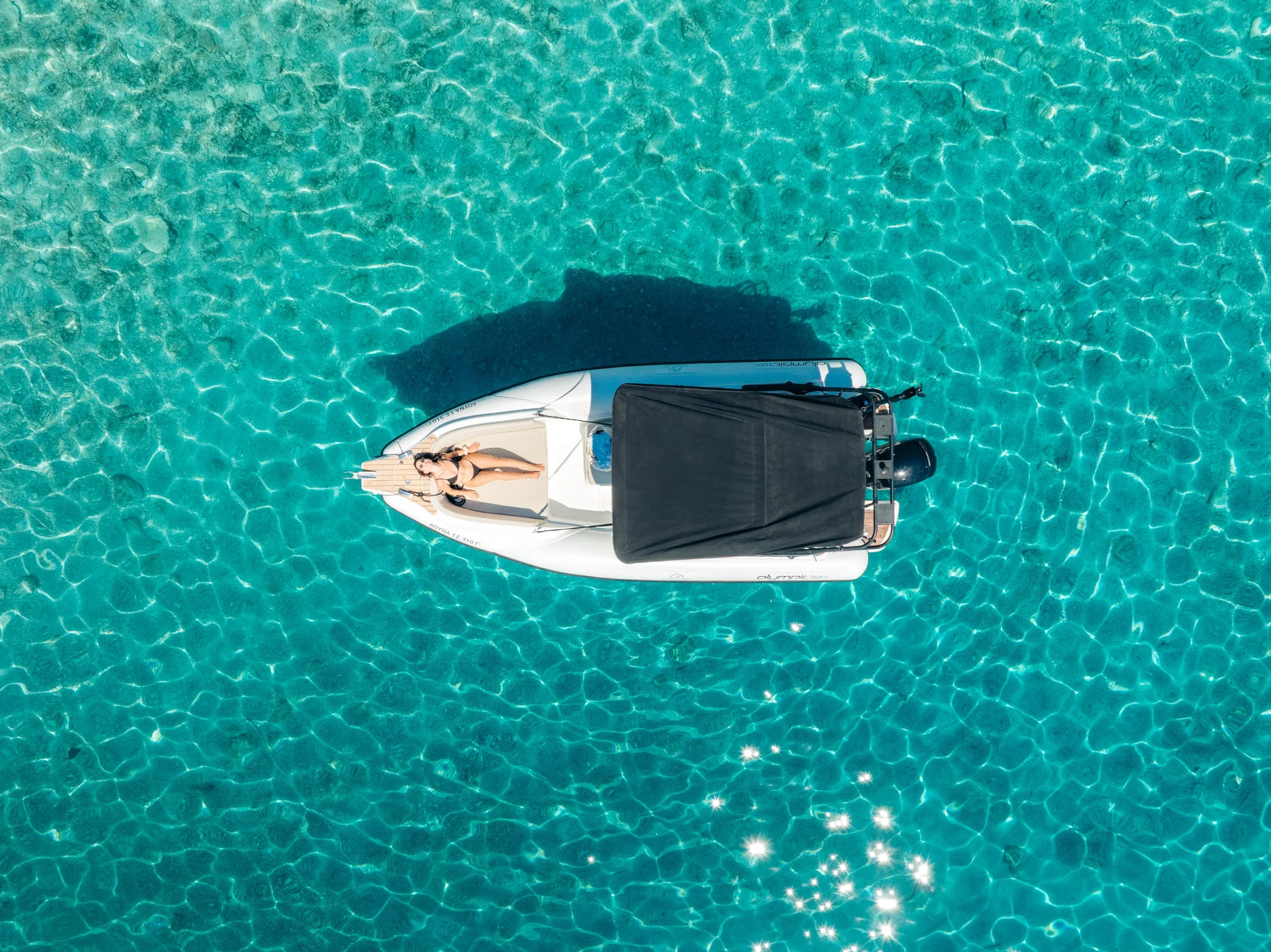 A woman in a black swimsuit lying on a small white boat on clear turquoise water, with a black cover over part of the boat.