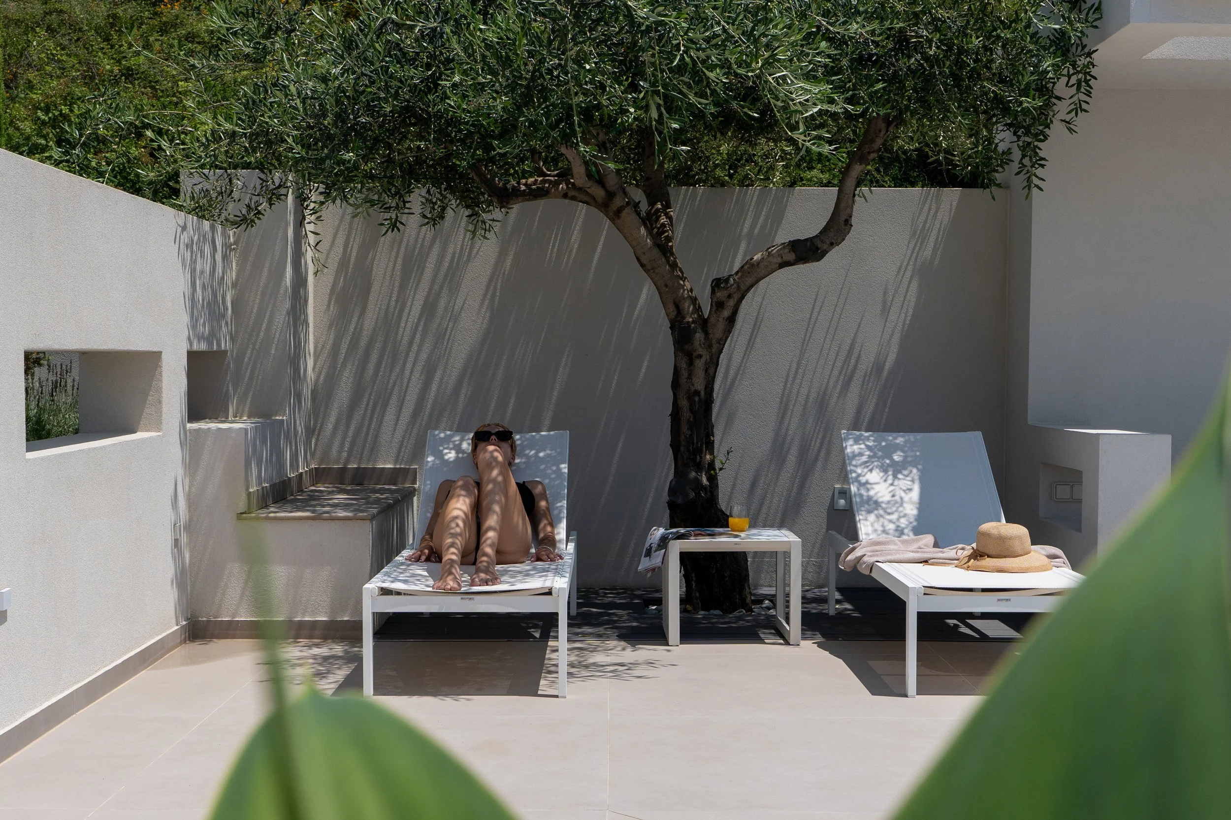 A woman lounging on a white chaise under a tree on a patio with white walls. There is a straw hat and a towel on the right chaise and a glass of orange juice on a small table between the loungers.