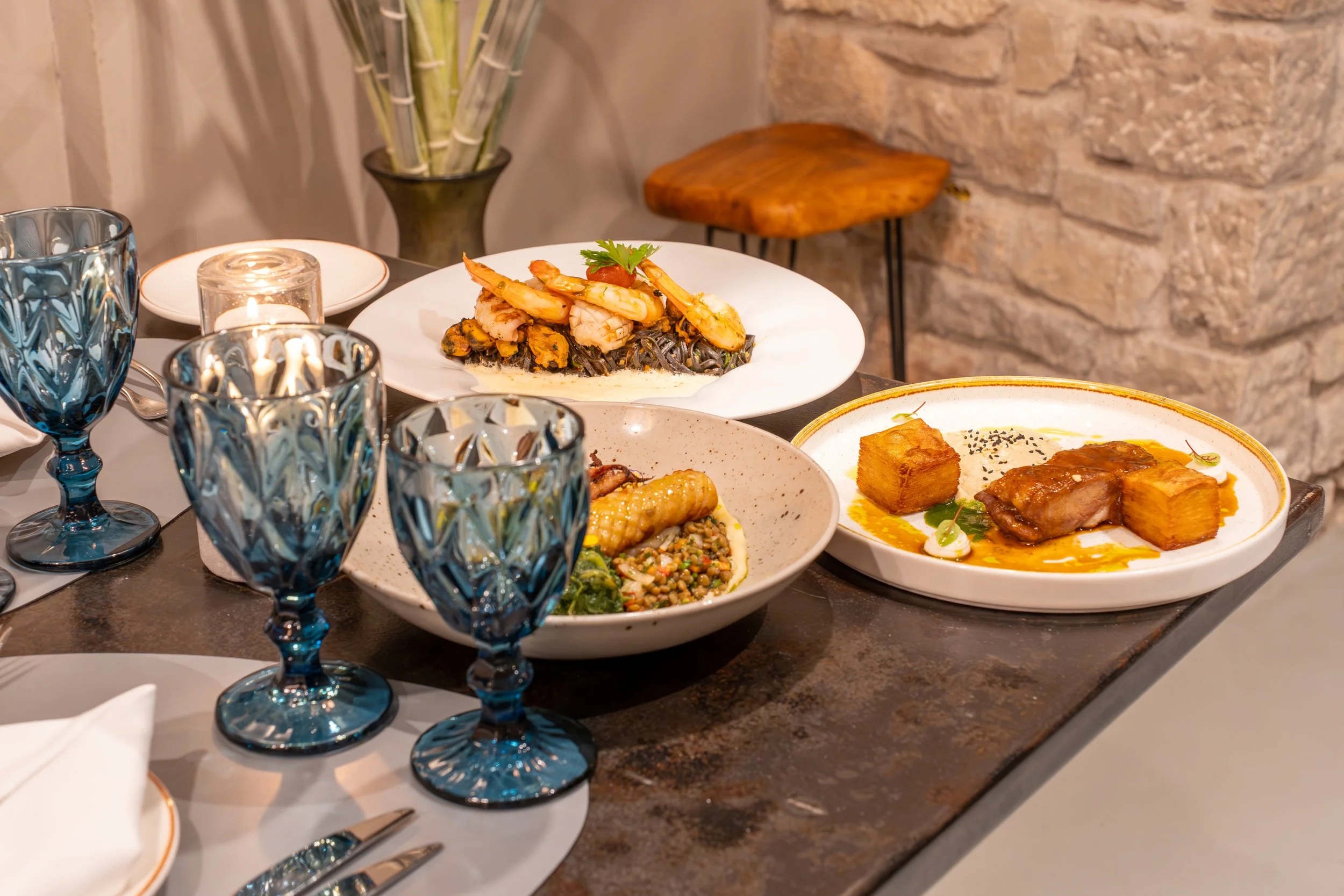 Elegant table with three blue glasses, three white plates with gourmet dishes, a lit candle, and a napkin with utensils, set against a stone wall background.