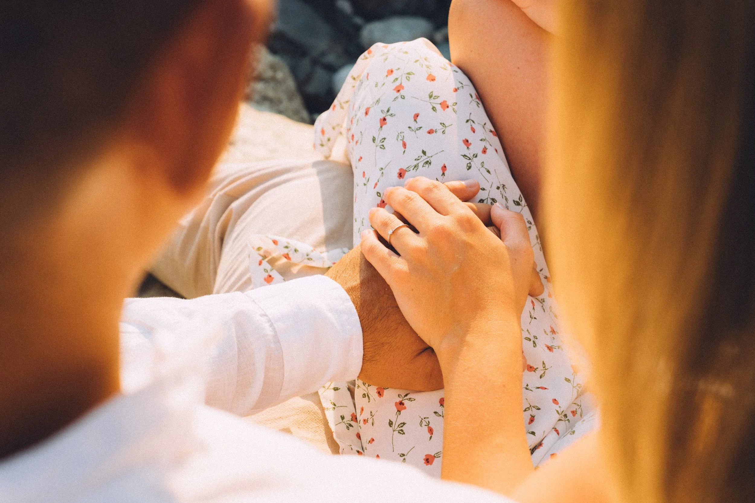 A couple holding hands on a woman's pregnant belly, with the woman wearing floral pants and another person wearing a white shirt, outdoors in natural lighting.