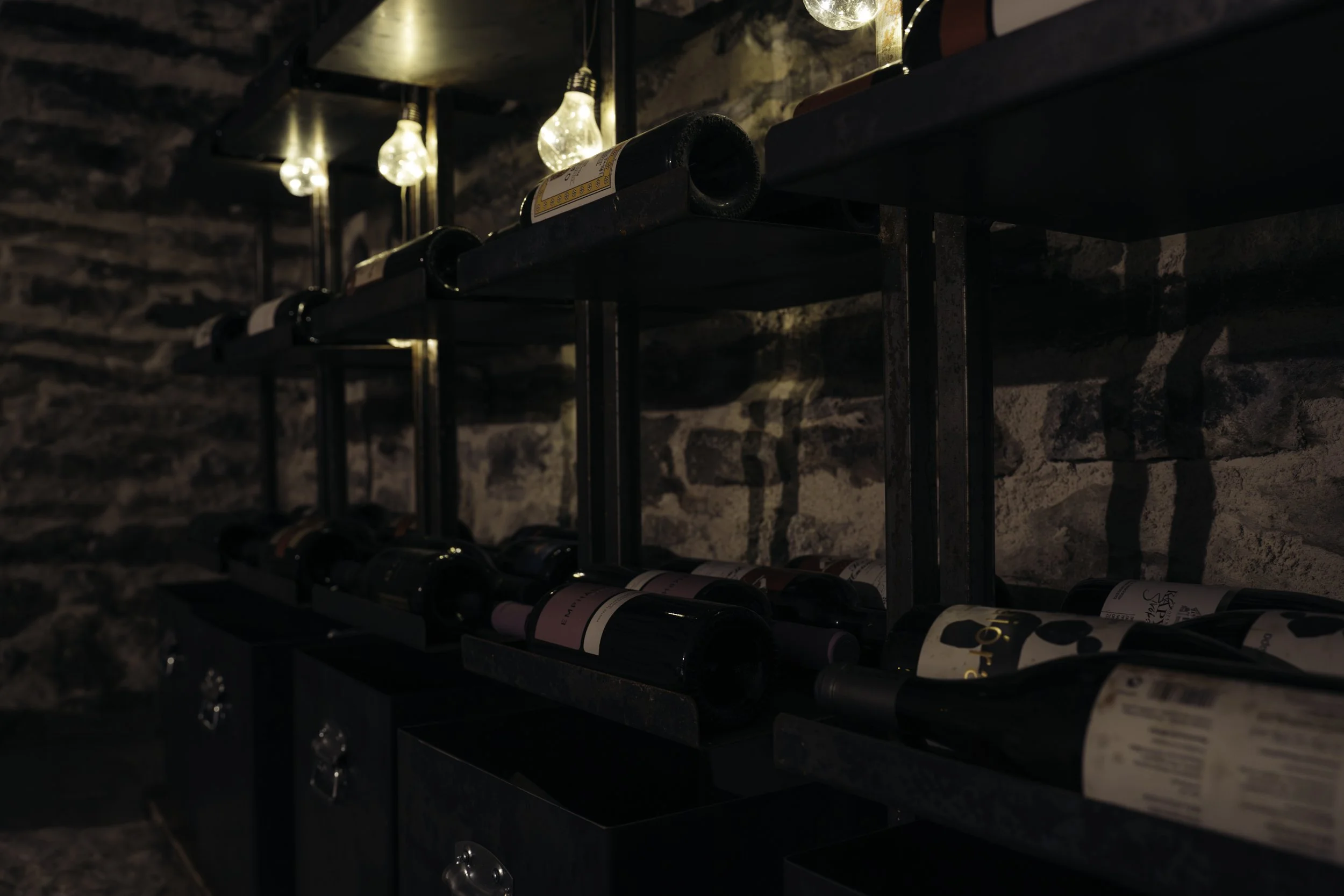 A wine rack against a brick wall with several bottles of wine stored horizontally, illuminated by warm hanging light bulbs.
