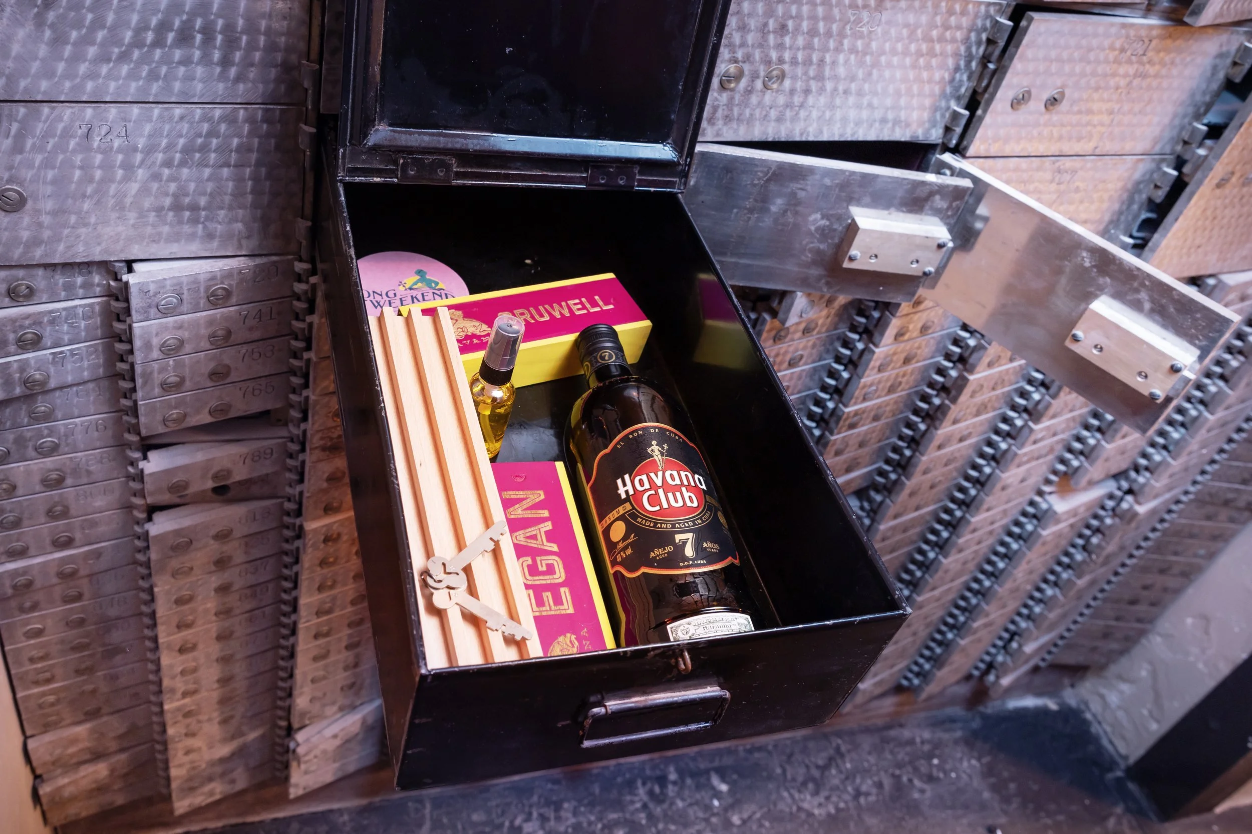 An open metal mailbox containing a bottle of Havana Club rum, a small bottle of what appears to be whisky, a box of matches labeled 'BRUWELL,' a pack of wooden sticks, and a circular sticker that says 'LONG WEEKEND.'