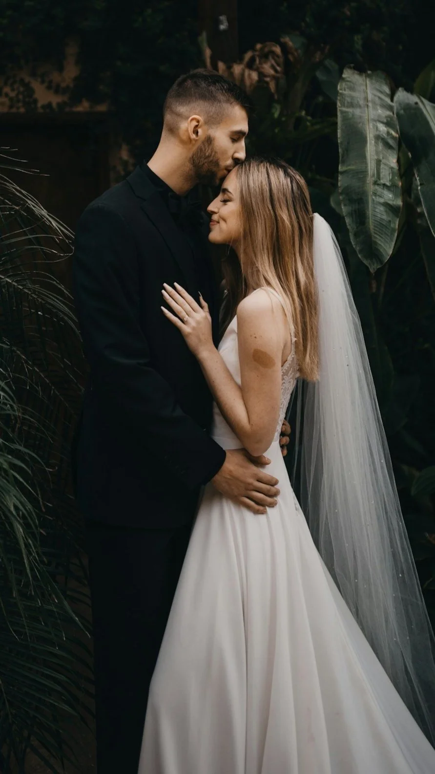 Bride and groom embracing, bride in white gown and veil, groom in black suit, surrounded by greenery.
