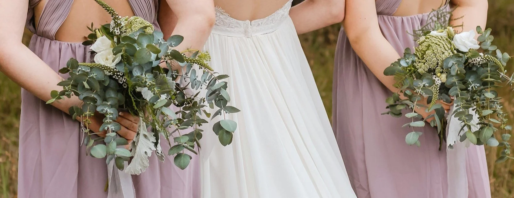 Bride in white dress and bridesmaids in lavender dresses holding bouquets of greenery.