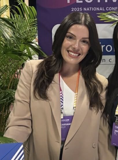 A person with pink nail polish holding a smartphone at a wooden desk with a laptop, TV remote, and car key nearby.