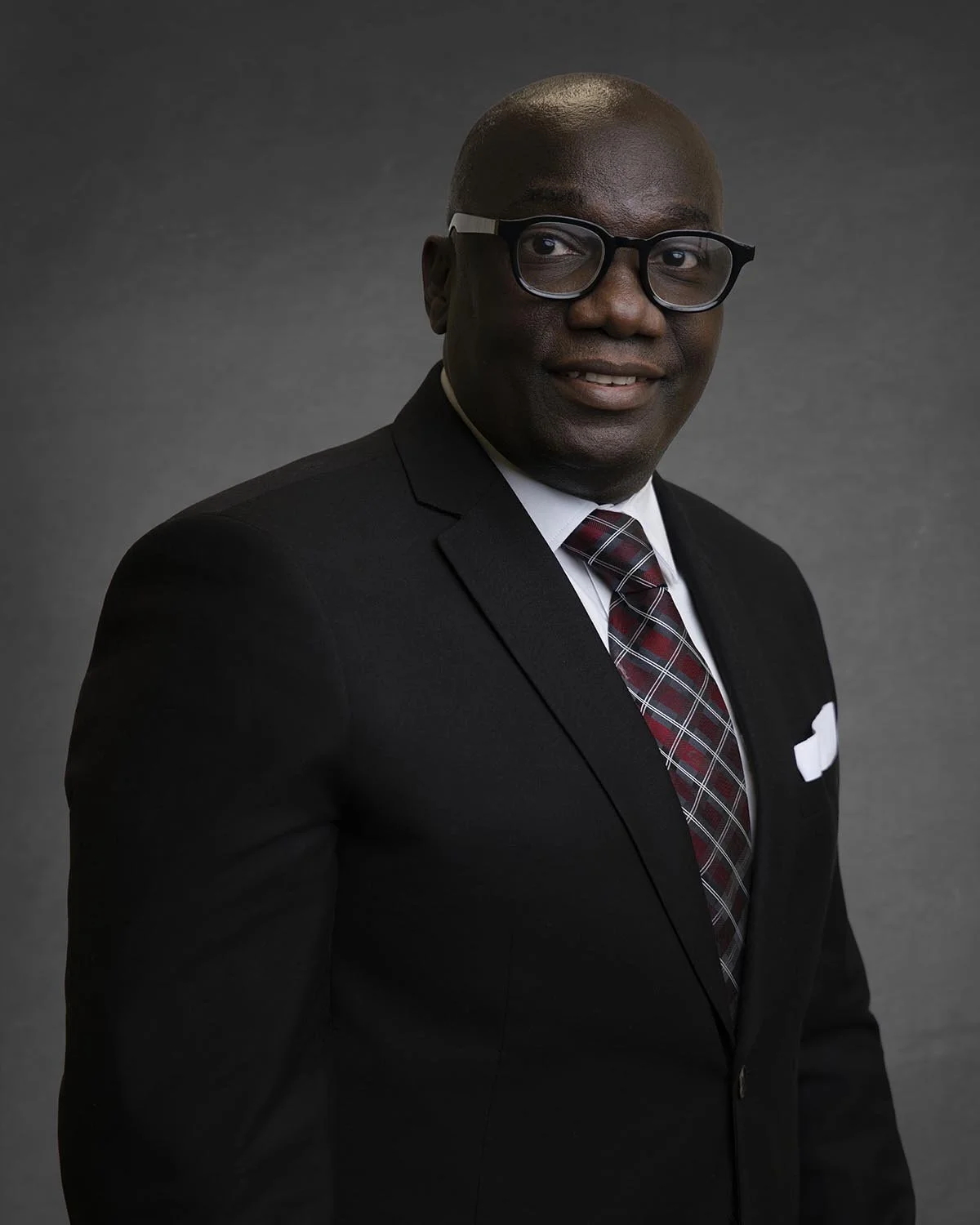 Man wearing glasses, a dark suit, and a red tie with a matching pocket square, posing against a dark background.
