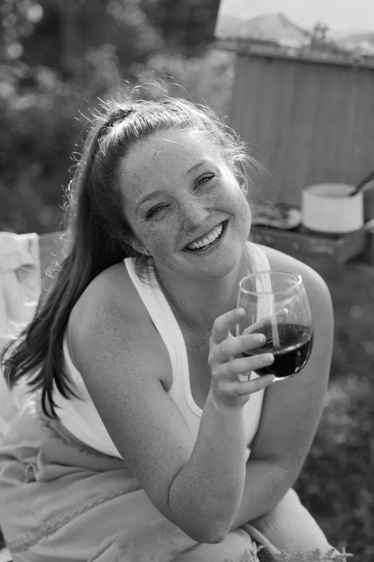 black and white image of the artist Ashley Hendrix cheers-ing the camera with a glass of wine and an easel behind her in a field