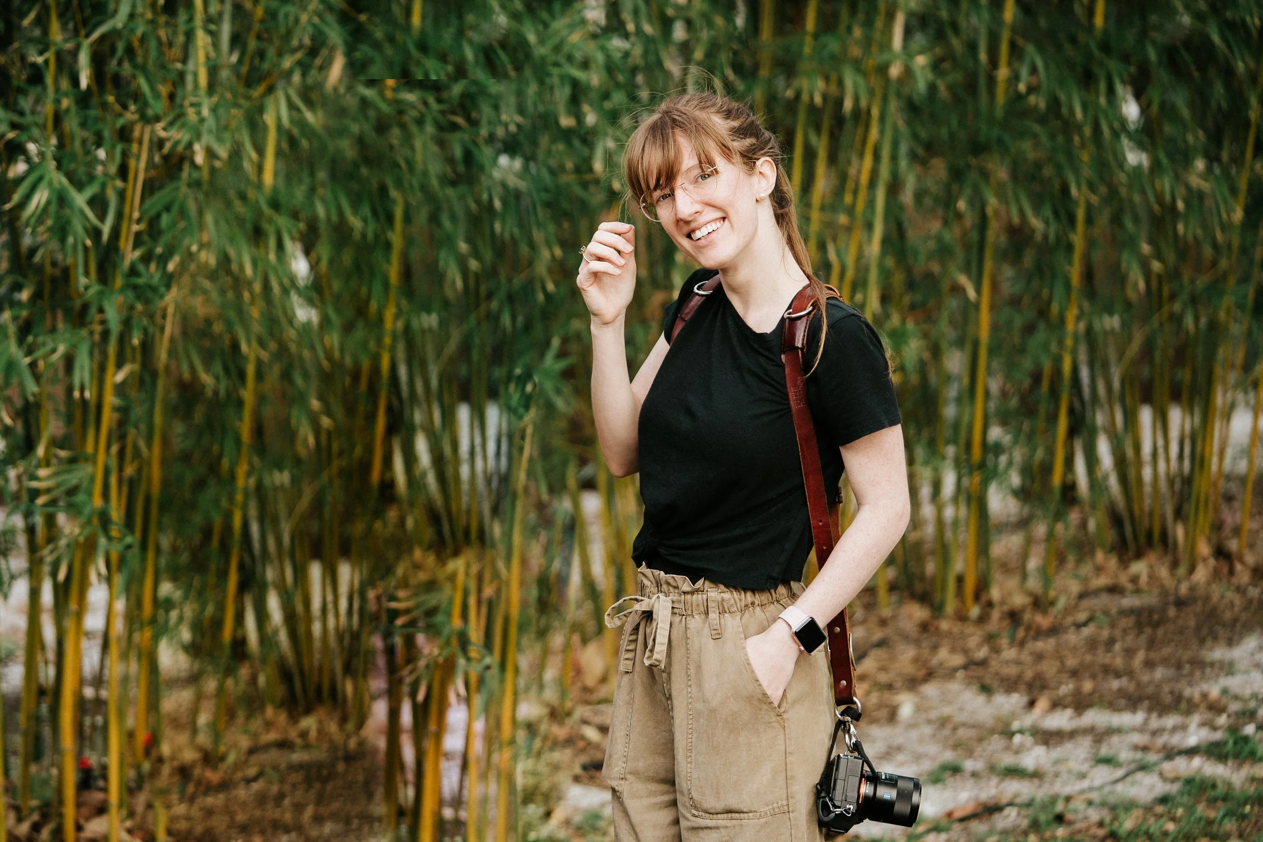 A young woman smiling outdoors with a camera hanging from her waist, standing in front of lush green bamboo.