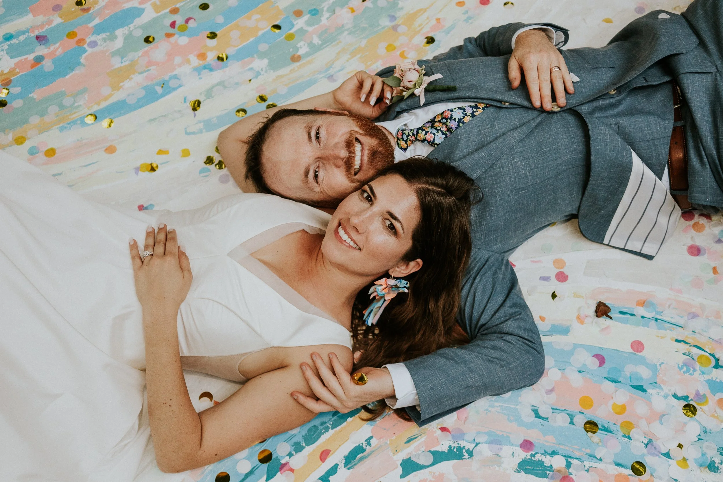 A happy couple lying on the floor, surrounded by colorful confetti and torn paper streamers, smiling at the camera. The man has a beard and is wearing a gray suit with a floral tie, and the woman has long dark hair and is wearing a white dress and floral earrings.