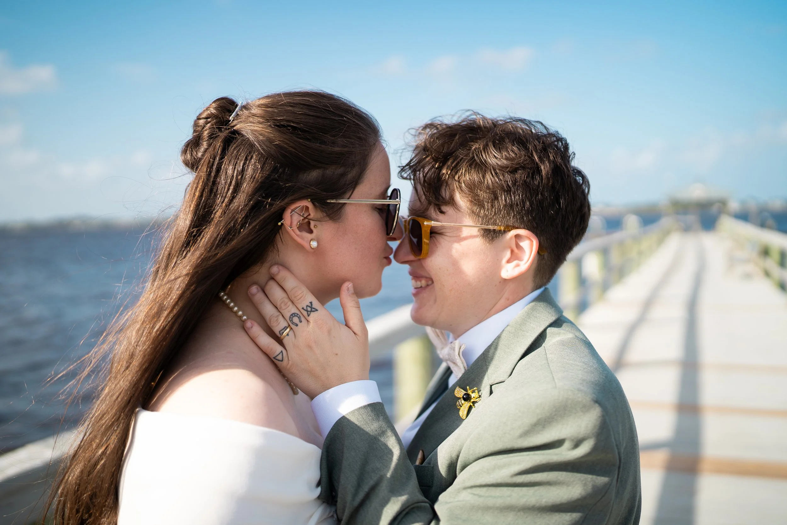 A couple dressed in wedding attire sharing a kiss on a marina dock, with water and sky in the background.