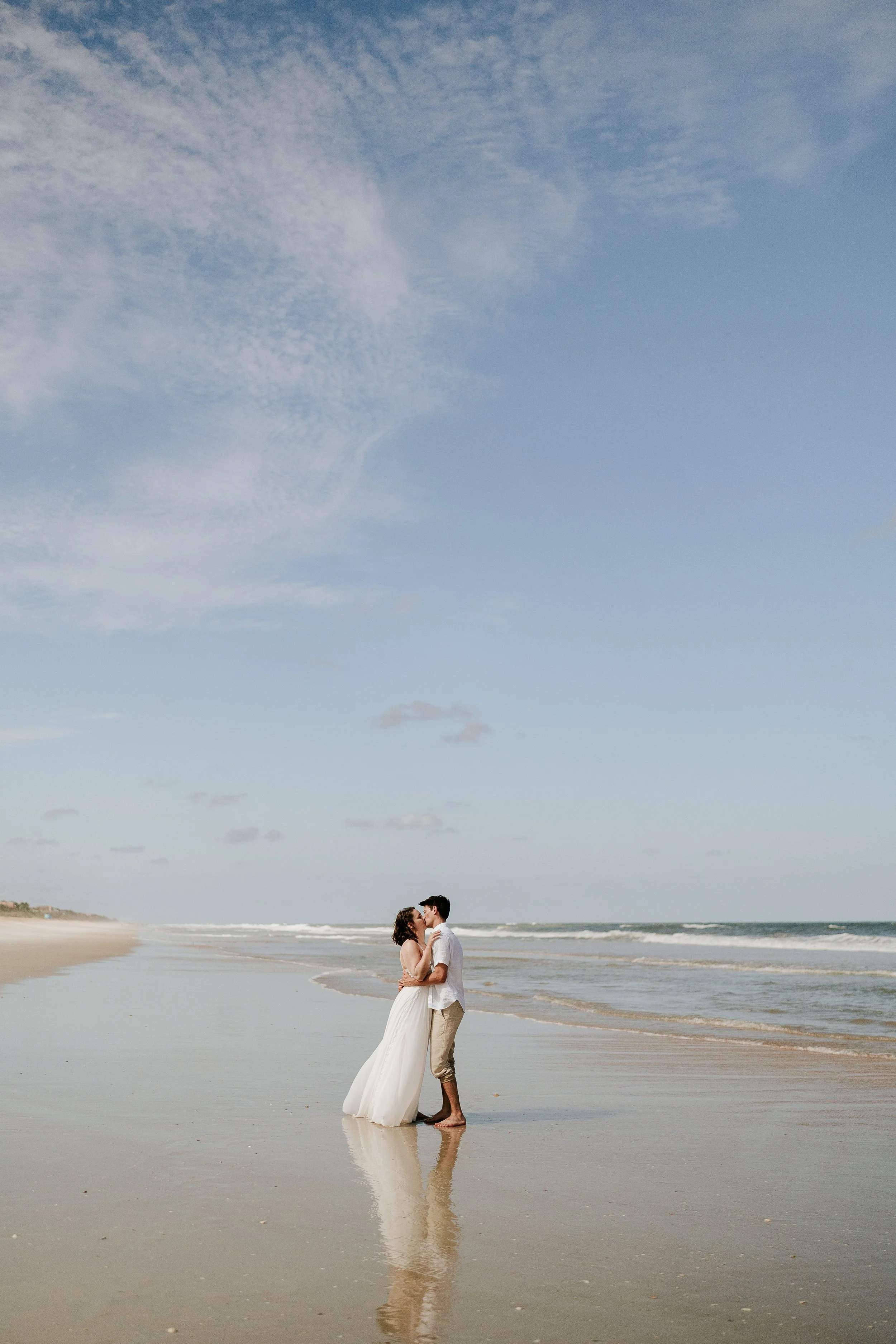 A couple kissing on the beach, with the woman in a white wedding dress and the man in casual attire, standing in shallow water under a partly cloudy sky.