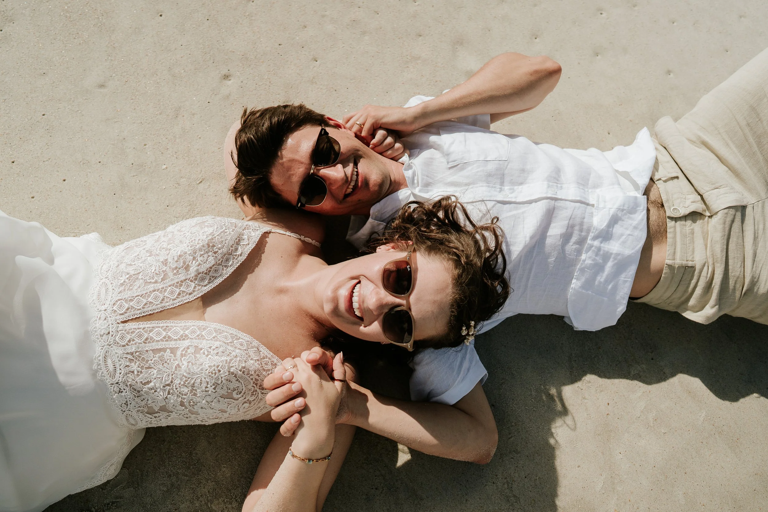 A man and a woman lie on the sand, smiling and holding hands, both wearing sunglasses and casual summer clothing.