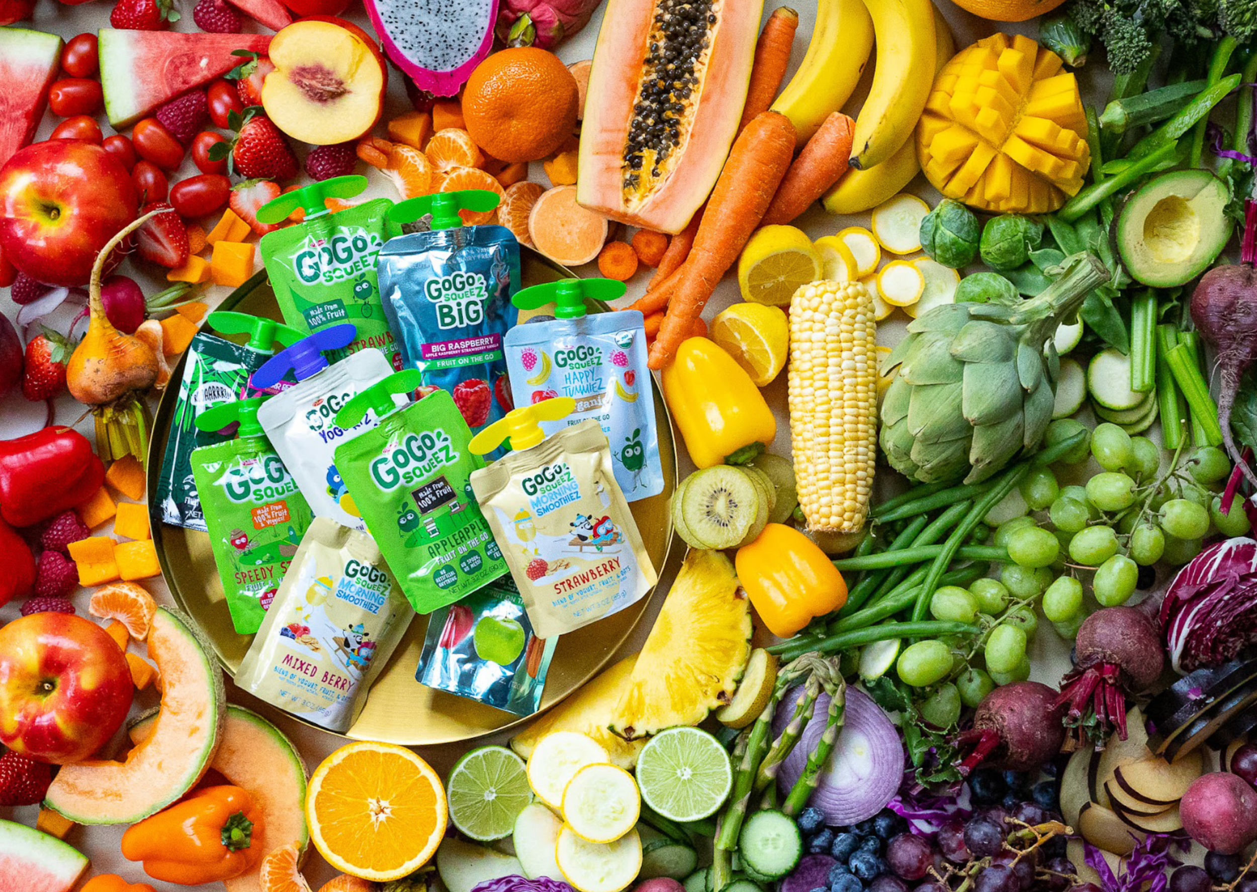 Colorful array of fresh fruits and vegetables surrounding a tray of Gogo SqueeZ fruit pouches.