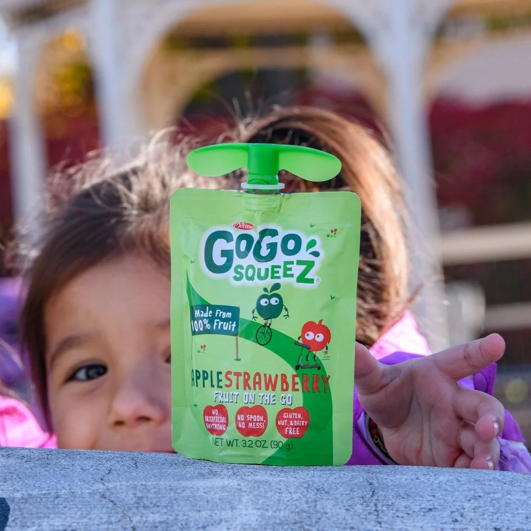 A young girl holds a green pouch of Gogo Squeeze alternative fruit snack with a superhero design, labeled 'Applestrawberry', outdoors with sunlight.