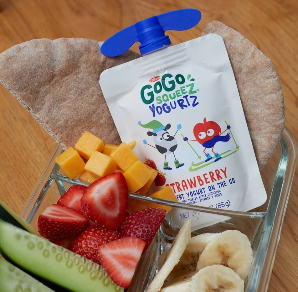 A clear container with sliced strawberries, banana slices, cubed mango, cucumber slices, and a go-go squeeze yogurt pouch with a blue cap inside a paper wrapper.