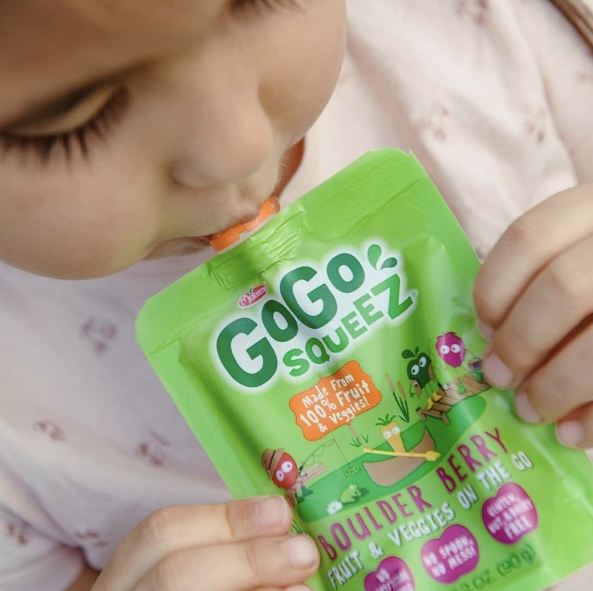 A young child drinking from a green pouch labeled 'GOGO SQUEEZ' with a flavor of 'Boulder Berry' and a note that it is made from 100% fruit and veggies.