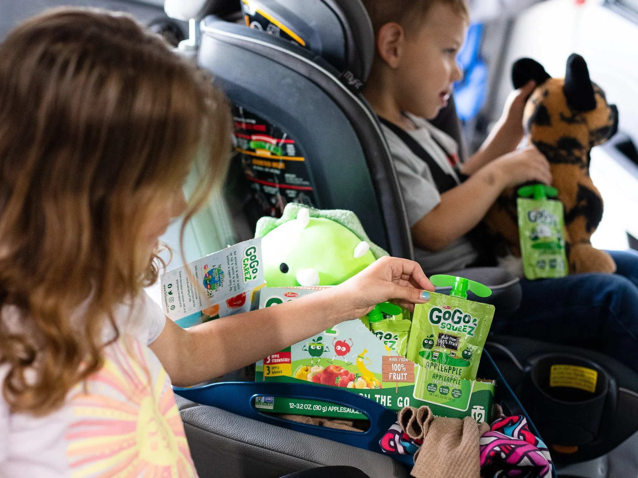 Two children sitting in car seats during a car ride, one girl with curly red hair reaching into a tray of Gogo SqueeZ fruit snacks, the other girl with straight hair holding a stuffed animal.