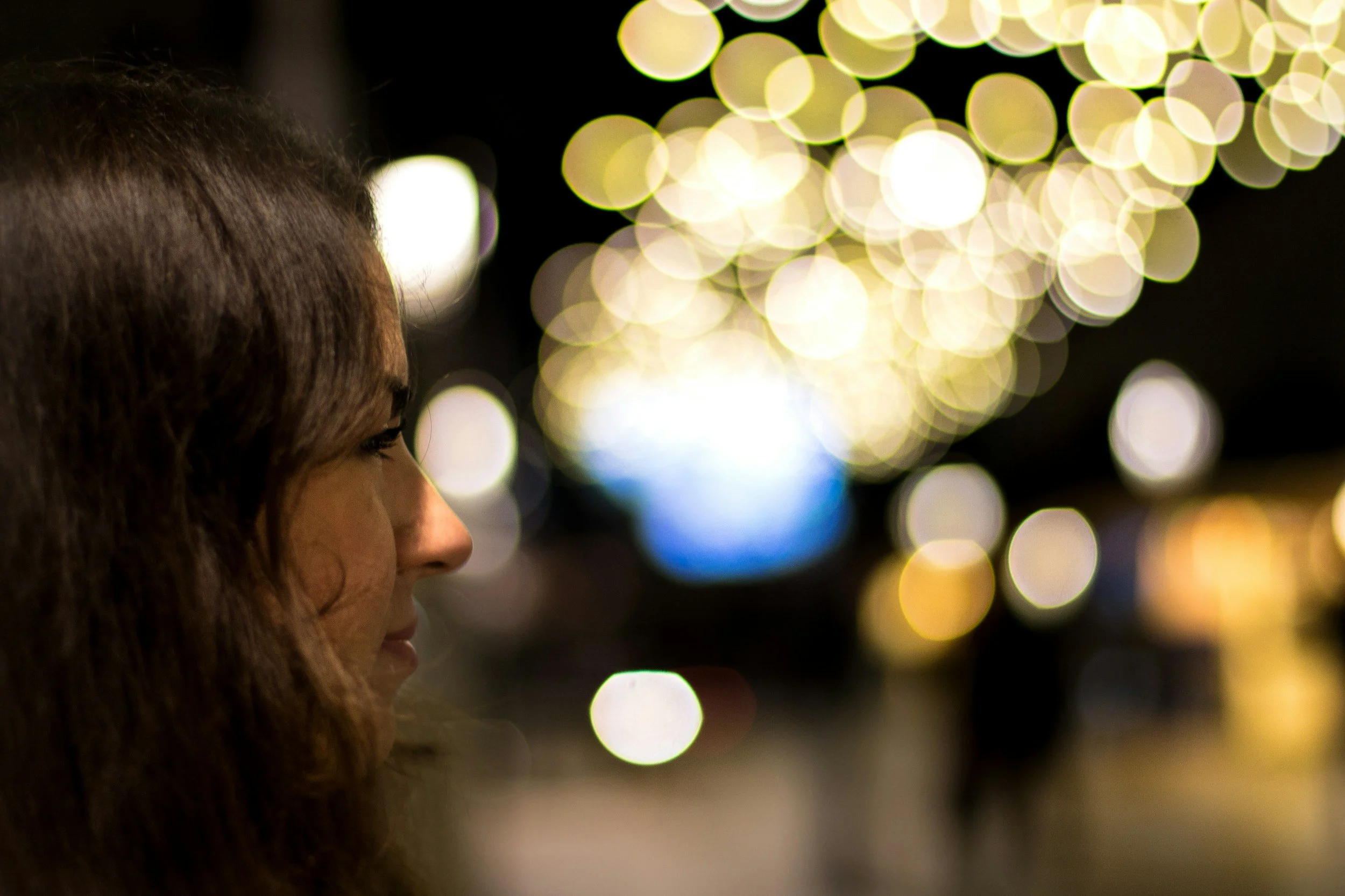 Close-up of a woman's face with dark hair, with a background of yellow bokeh lights at night.