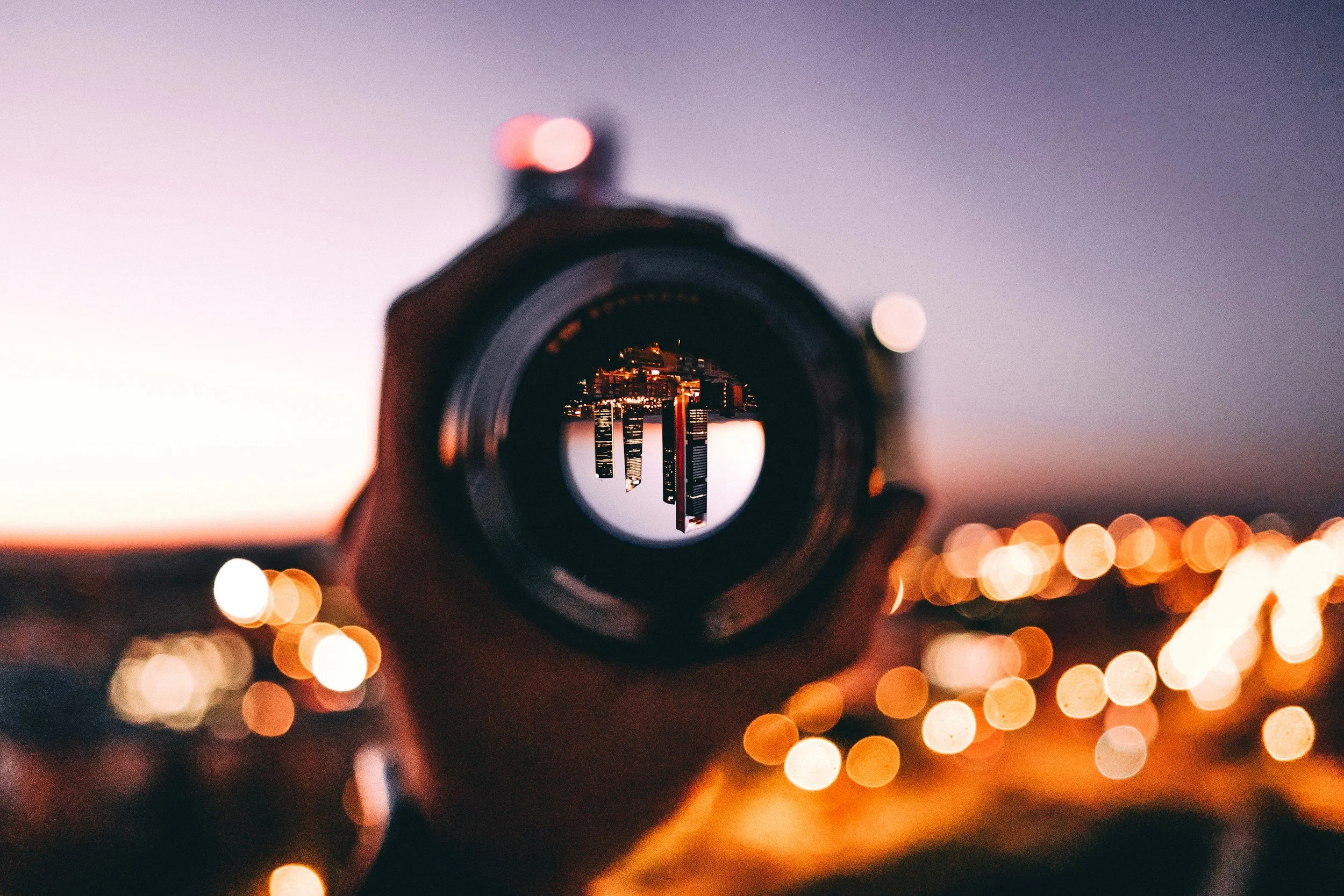 A camera lens reflecting city skyscrapers during sunset or twilight, with blurred orange street lights in the foreground.