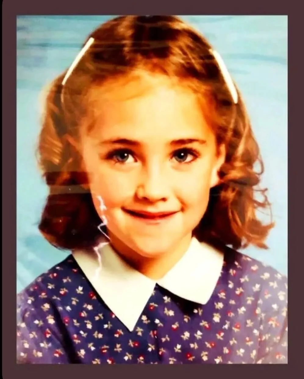 A young girl with curly red hair and blue eyes, wearing a navy dress with a white collar, smiles at the camera. The background features a blue sky or water.