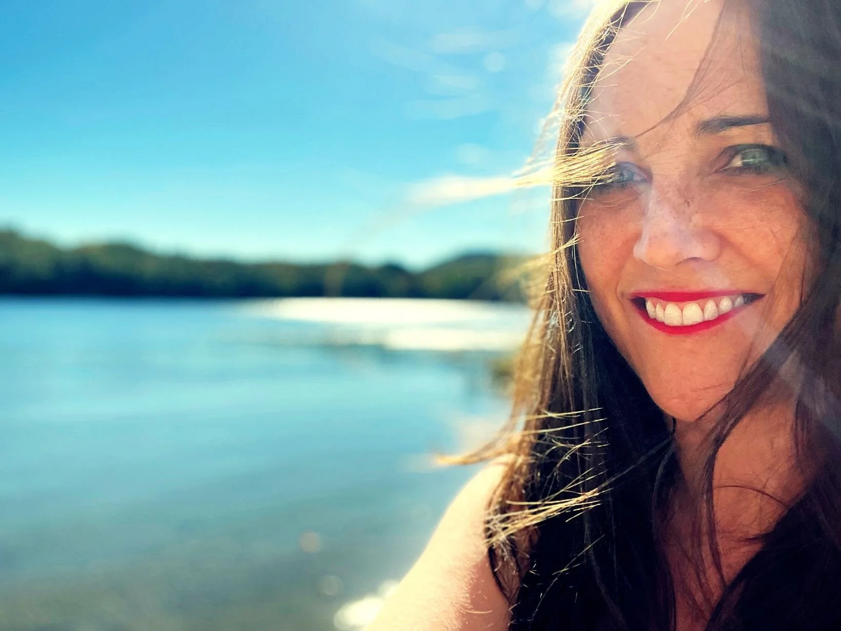 A close-up of a smiling woman with long dark hair outdoors near water under a clear blue sky.