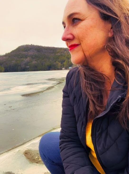 A woman with long brown hair and red lipstick looking to the right, sitting outdoors near a body of water with a hill in the background.