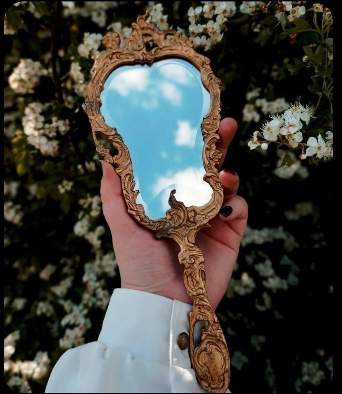 A hand holding an ornate wooden hand mirror reflects a blue sky with clouds, with white blooming flowers in the background.