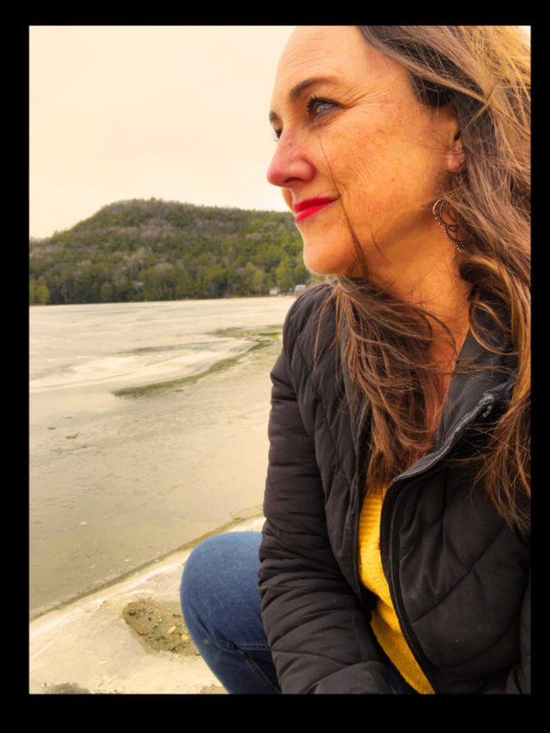 A woman with light skin and long brown hair sitting outdoors near a body of water with a green, forested hill in the background, wearing a black jacket and a yellow sweater, looking to the right with a slight smile.