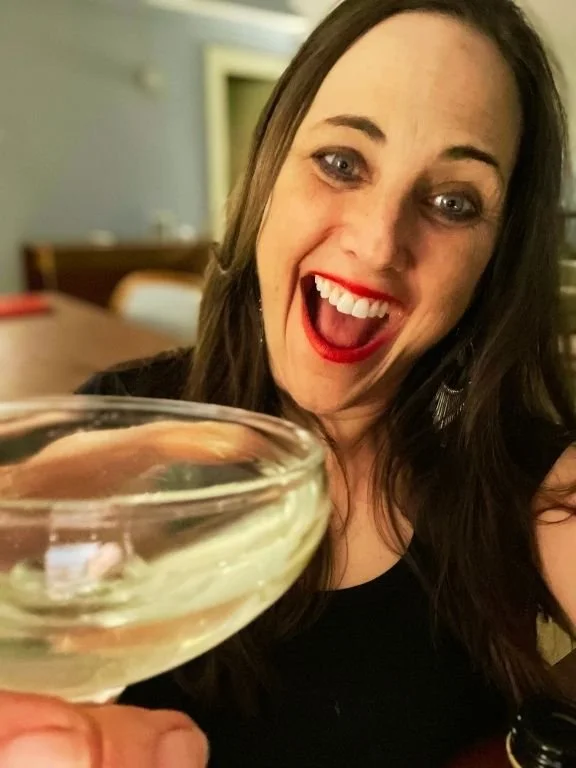 A woman with long dark hair, red lipstick, and earrings smiling and raising a glass in a celebratory toast in a cozy indoor setting.