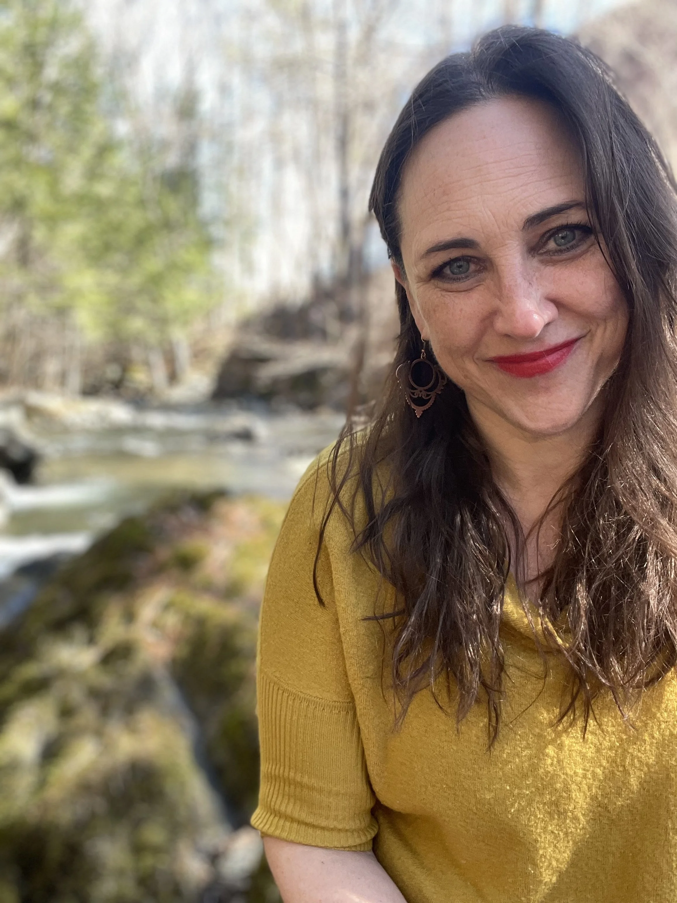 Close-up of a woman with long dark hair, blue eyes, red lipstick, wearing earrings and a yellow sweater, outdoors in a natural setting with trees and a stream in the background.