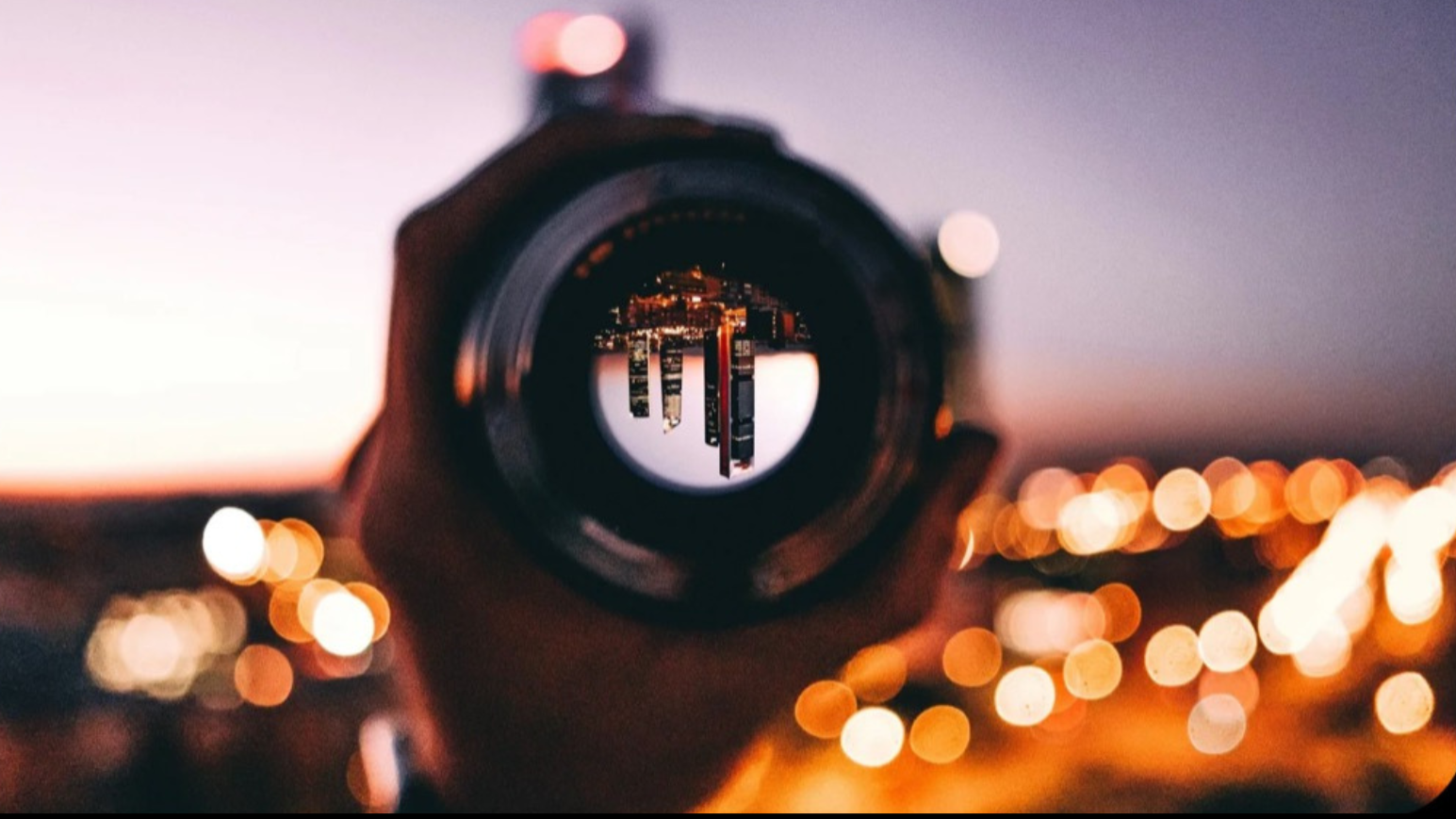 A camera lens reflecting a cityscape at sunset with city lights blurred in the background.