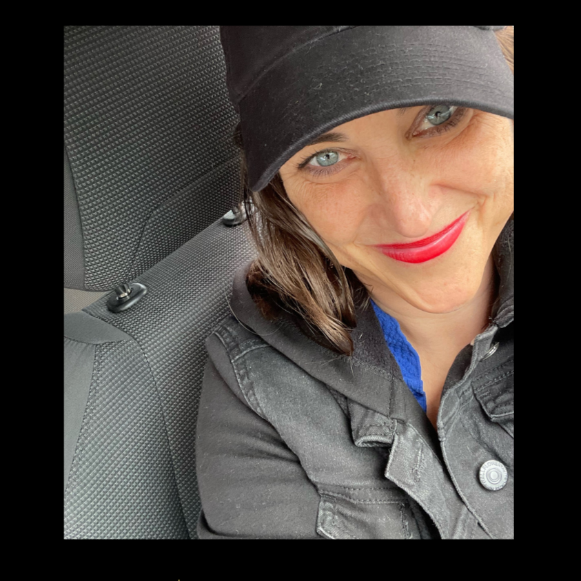 A smiling woman with bright red lipstick, blue eyes, and shoulder-length brown hair, wearing a black cap, black jacket, and blue shirt, sitting in a vehicle with a dark textured interior.