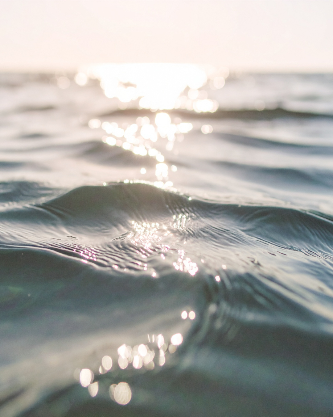 Close-up of ocean water with gentle waves and sunlight reflecting off the surface, with a bright sun over the horizon in the background.