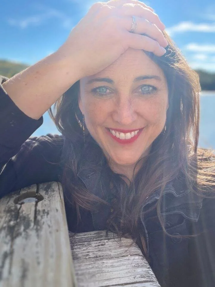 A smiling woman with blue eyes and brown hair outdoors, resting her head on her hand, with water and blue sky in the background.
