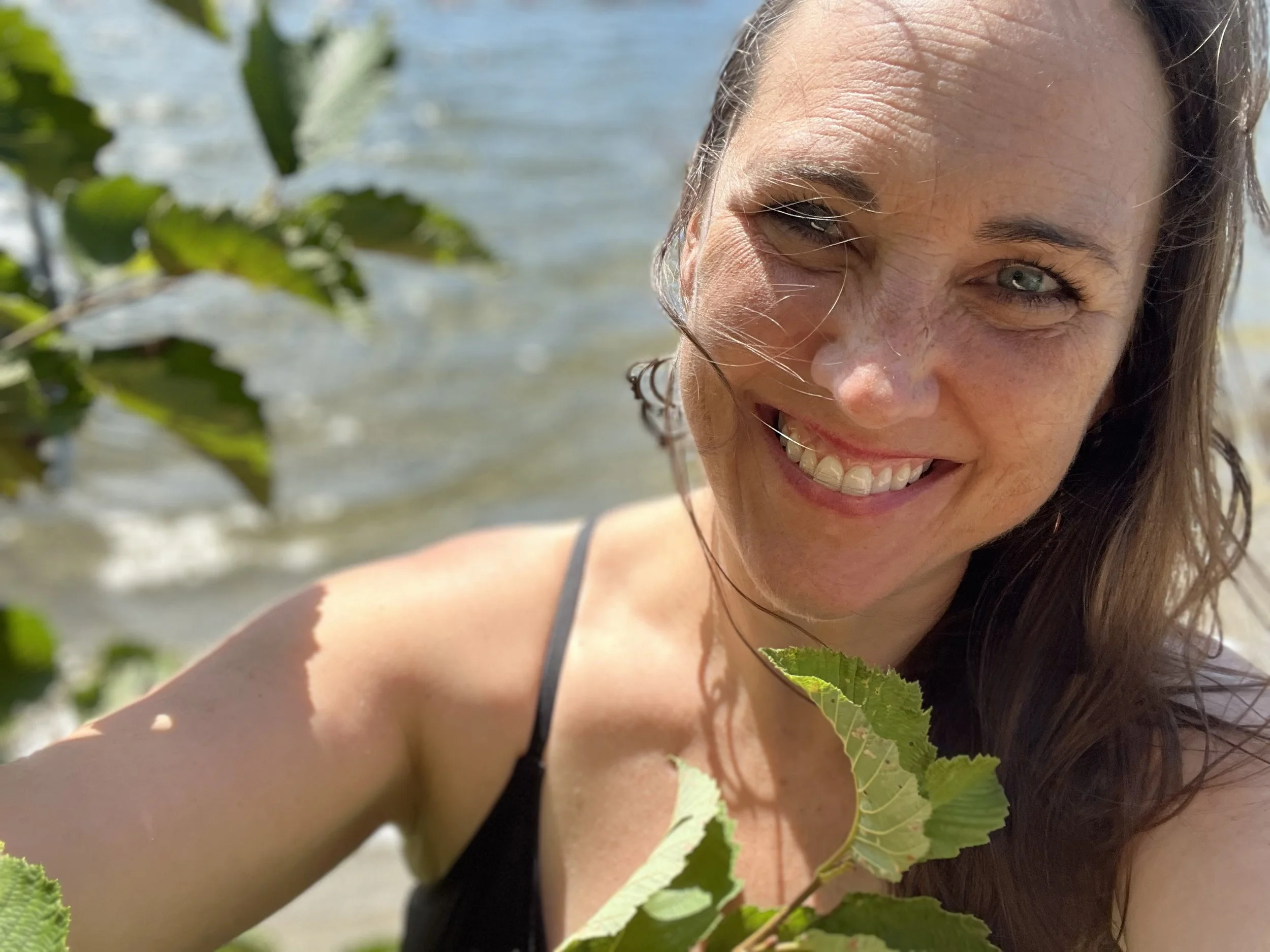 Close-up of a smiling woman with blue eyes and long brown hair, outdoors near water, with green leaves in the foreground.