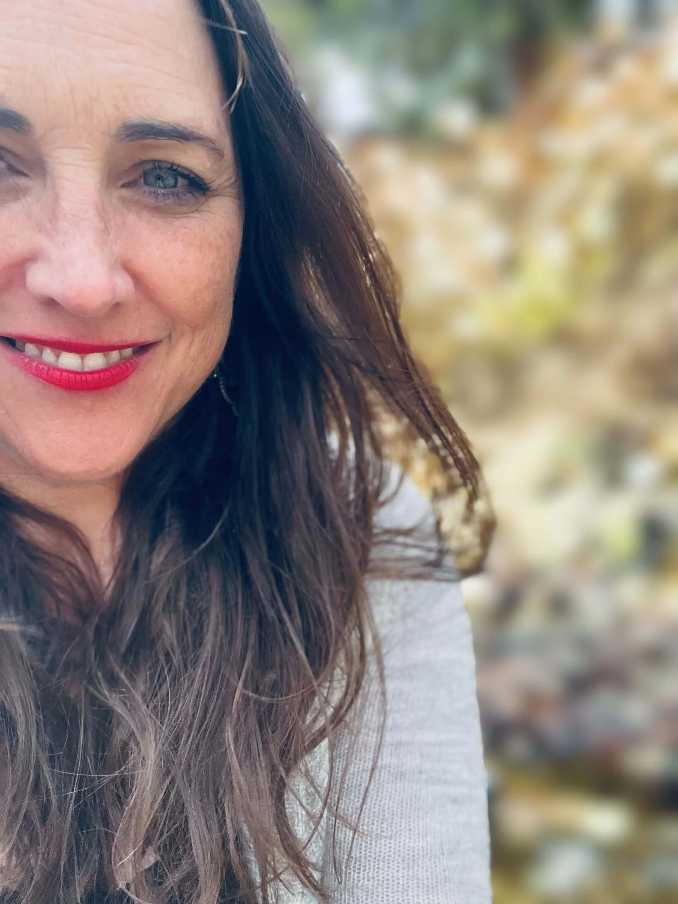 Close-up of a woman with brown hair and blue eyes smiling, wearing red lipstick, in front of a blurred outdoor background.