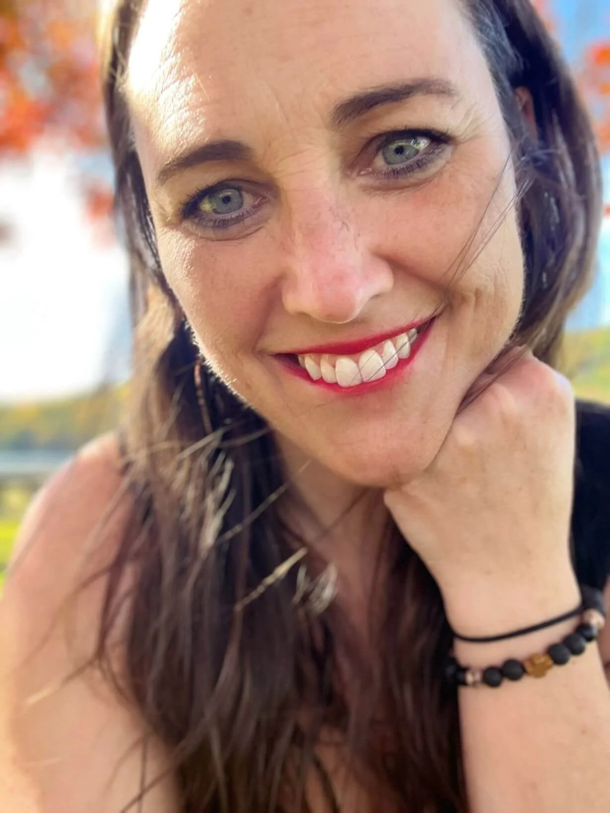 A woman with long brown hair and green eyes smiling outdoors during daytime, with trees and colorful leaves in the background.