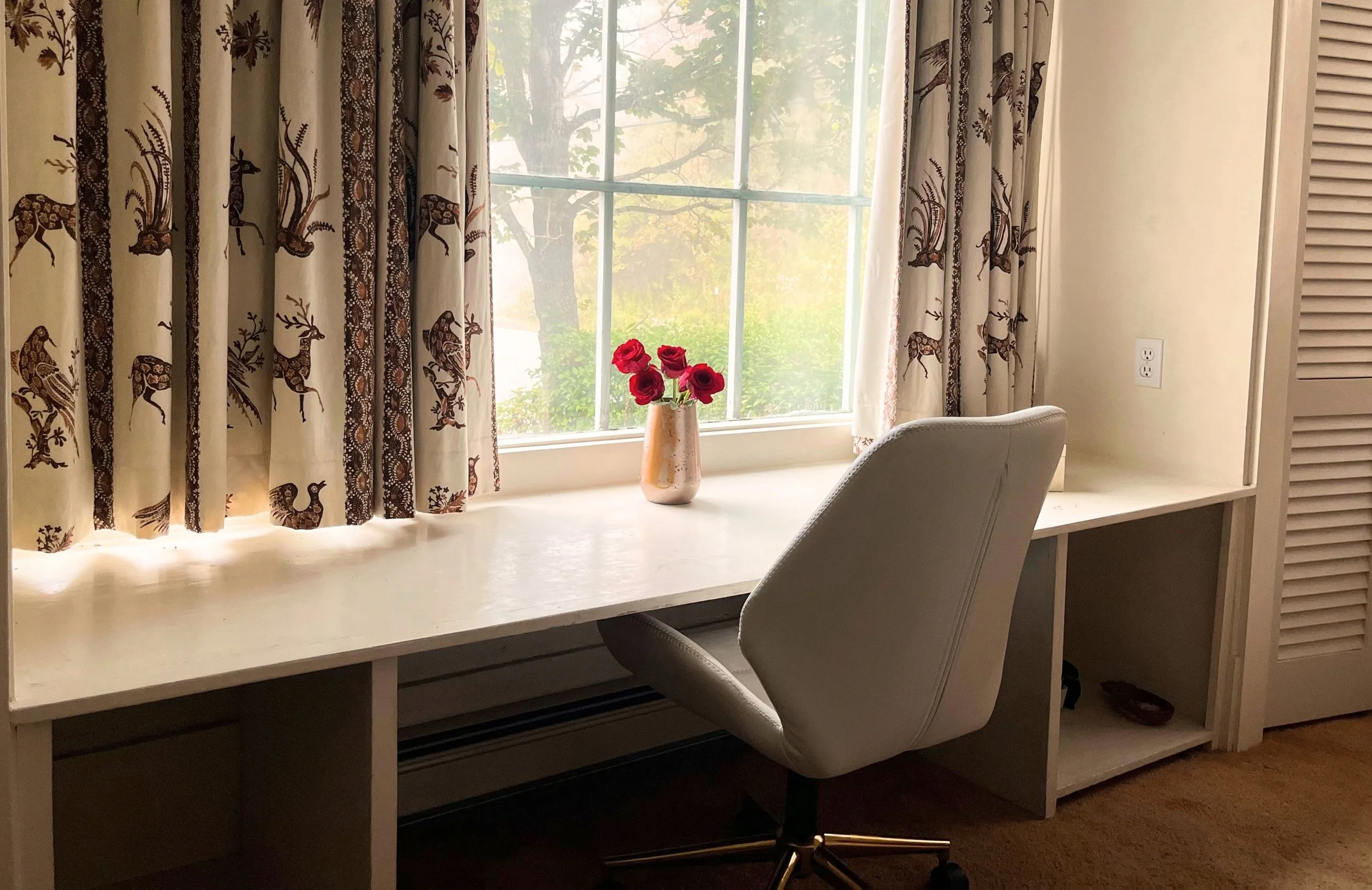 A study area with a white desk, a white office chair, and a window with patterned curtains featuring animals. There is a vase with red flowers on the desk, and greenery outside the window.