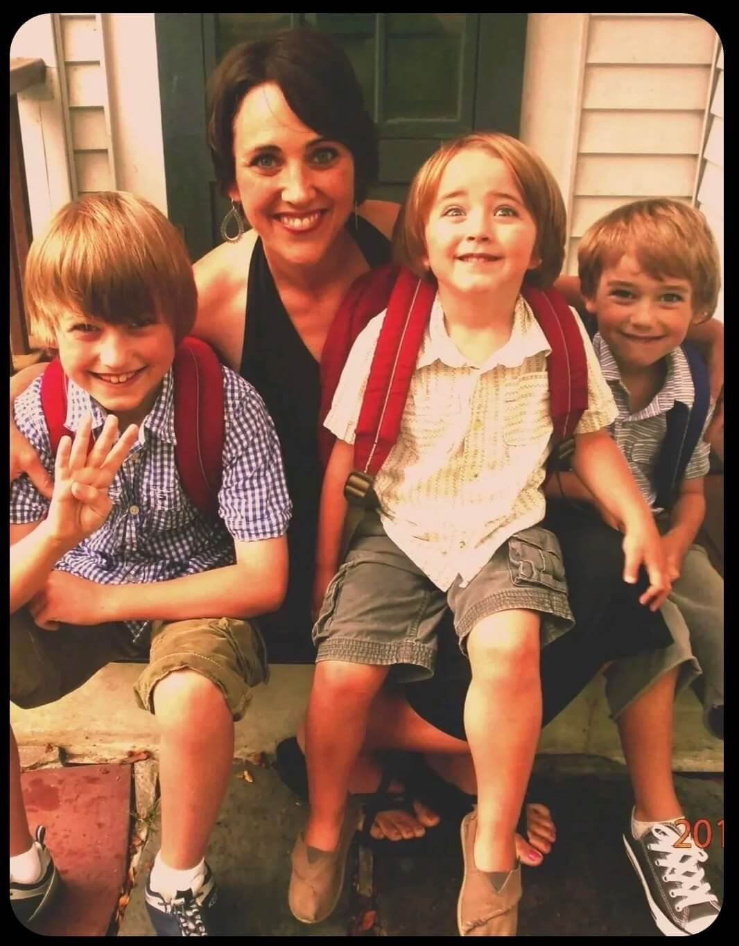 A woman with short dark hair and three young boys sitting on a porch. The boys are wearing backpacks, casual clothes, and sneakers. The woman and boys are smiling for the camera.